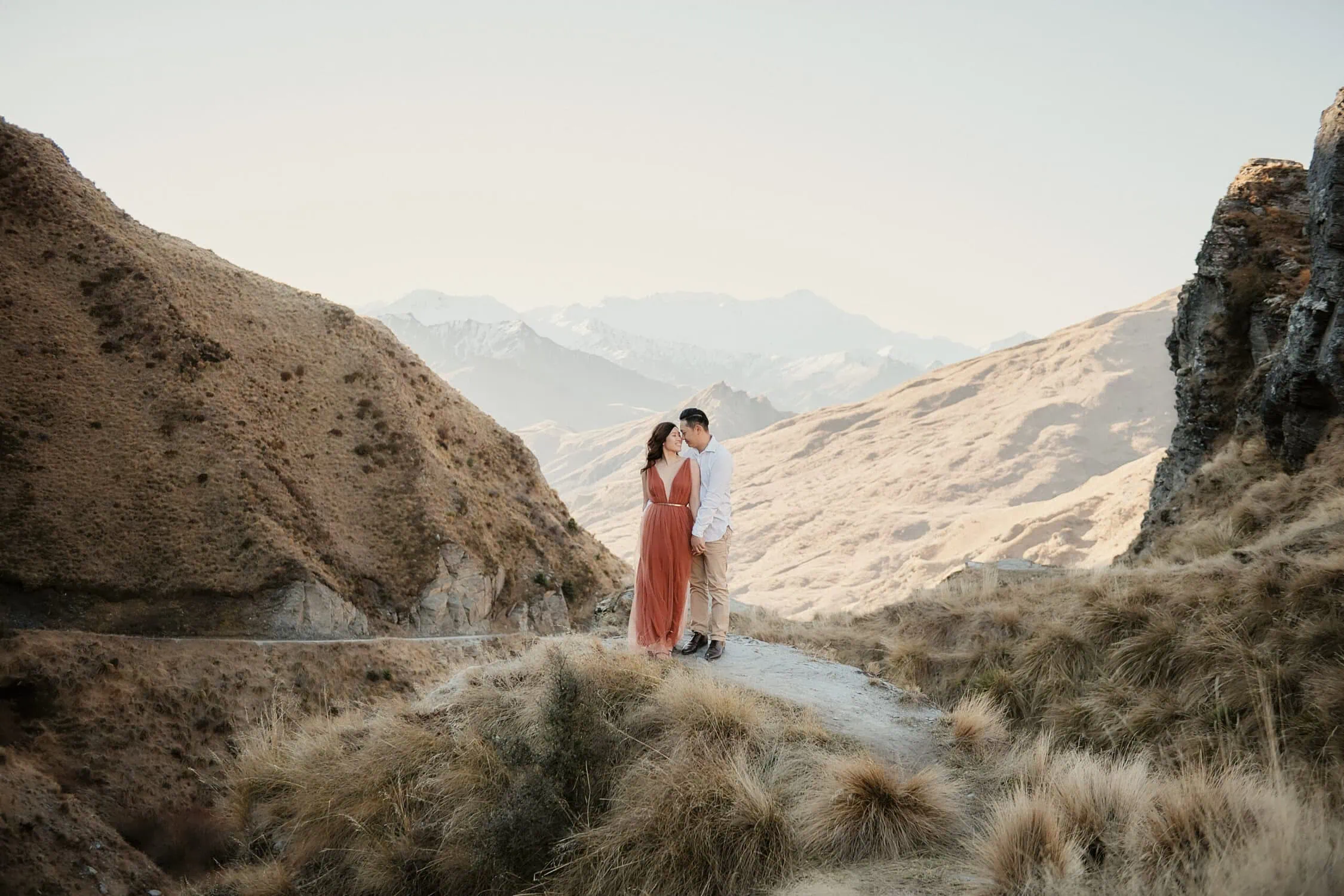 queenstown new zealand heli wedding elopement photographer videographer | A couple, Jennie & Eric, stands on a dirt path amidst the hilly terrain of Queenstown, NZ, with mountains visible in the background. The woman wears a long, rust-colored dress and the man wears a white shirt and beige pants—an idyllic scene for their pre-wedding shoot.