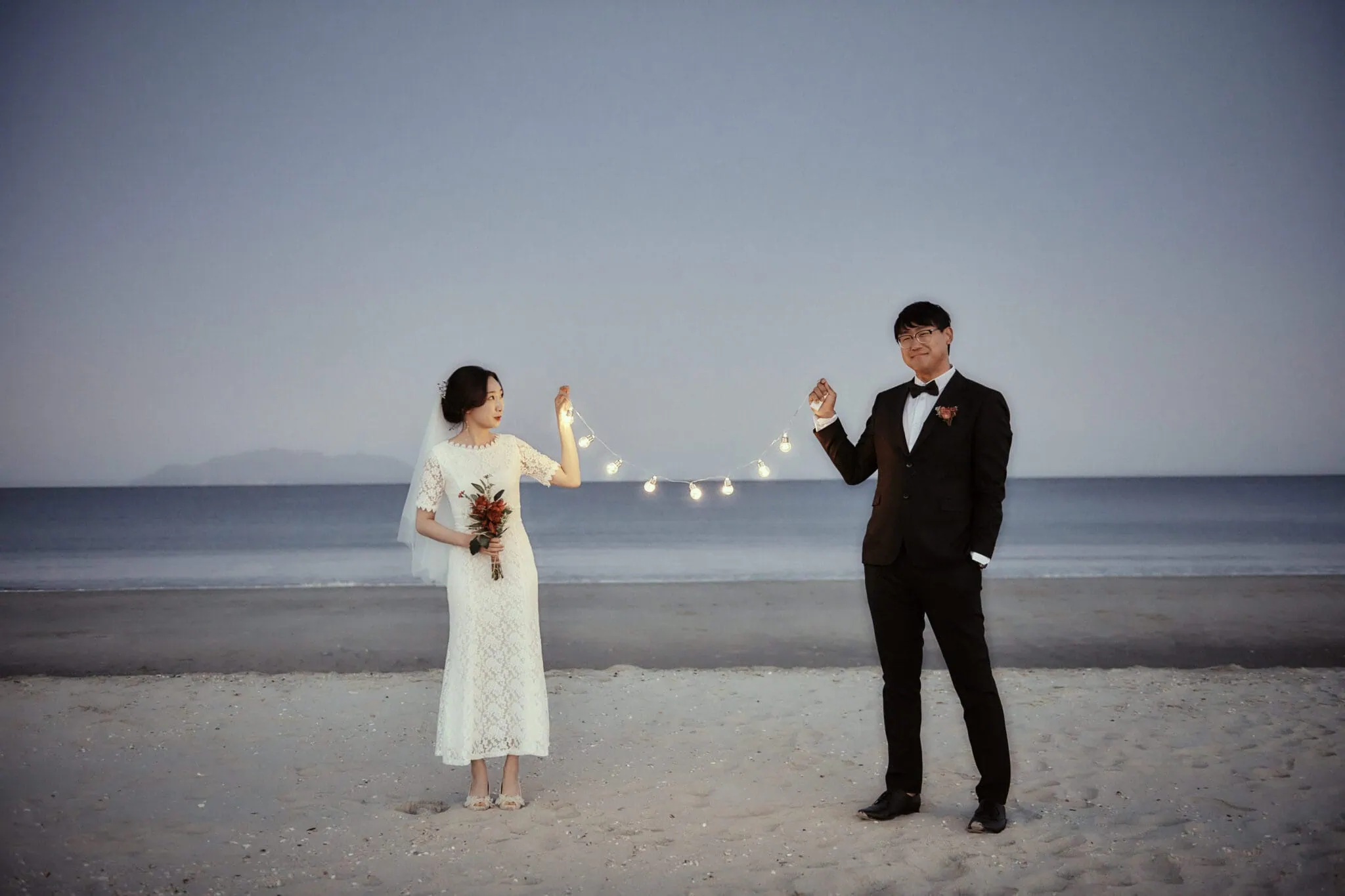 queenstown new zealand heli wedding elopement photographer videographer | A bride in a white dress and a groom in a black suit hold a string of lights between them while standing on a beach at dusk, capturing the magic of their NZ pre-wedding shoot.