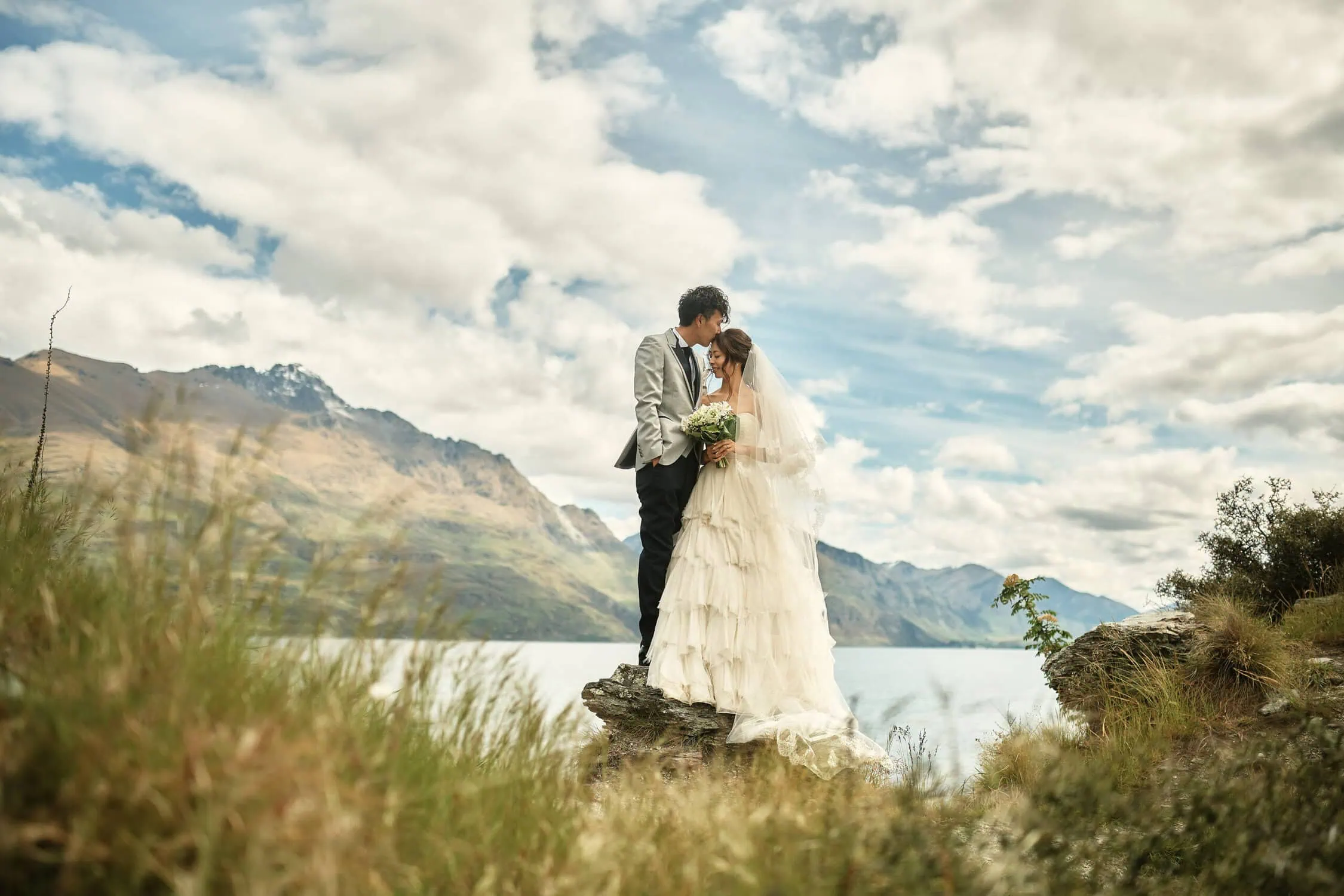 queenstown new zealand heli wedding elopement photographer videographer | A bride and groom stand on a rock by a lake in Queenstown, surrounded by mountains and tall grass, under a partially cloudy sky, capturing the perfect pre-wedding shoot.