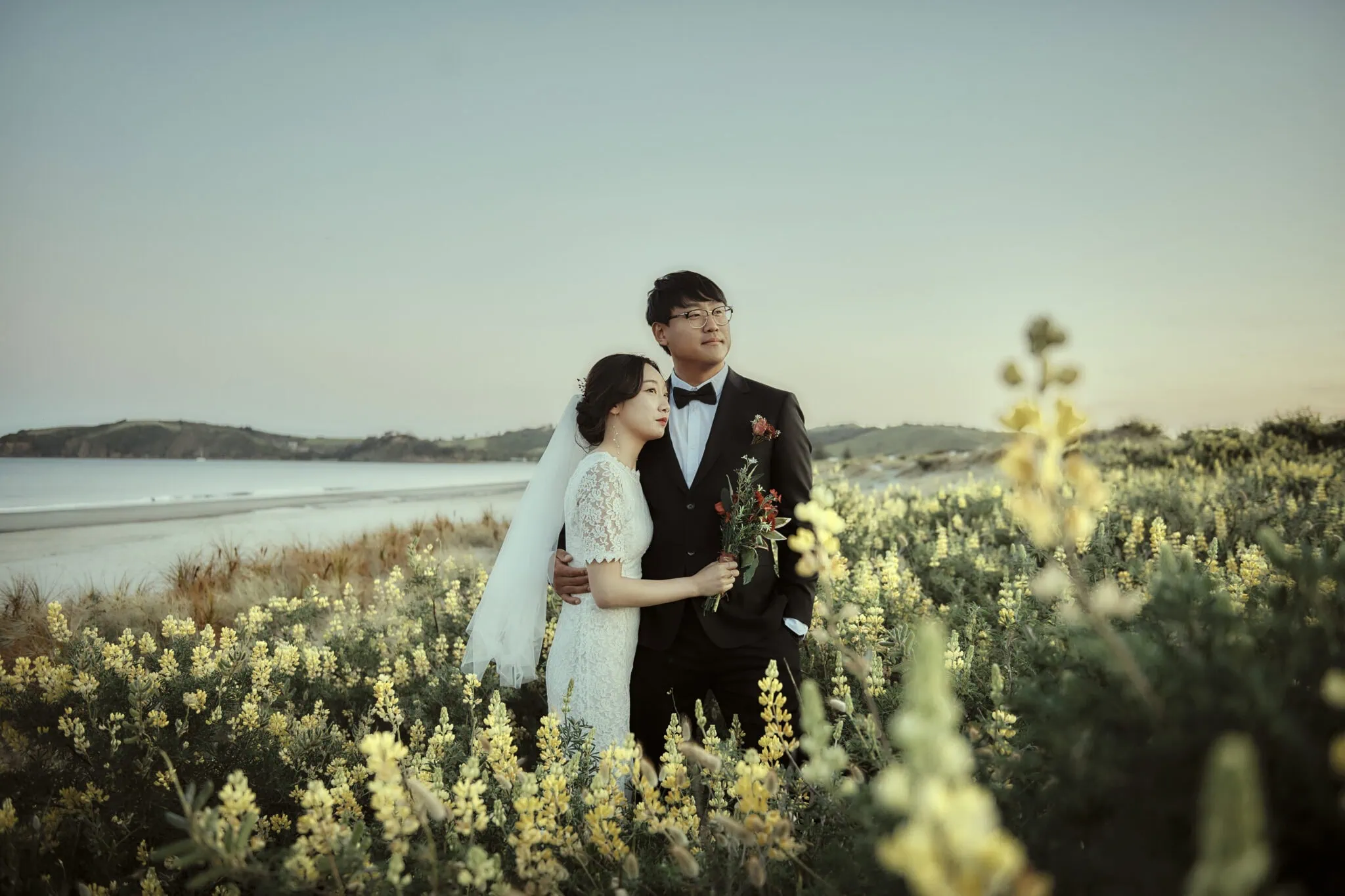queenstown new zealand heli wedding elopement photographer videographer | A bride and groom stand together in a field of yellow flowers by a beach, with the groom holding a bouquet of red flowers and the bride in a white dress and veil during their NZ Pre-Wedding Shoot.
