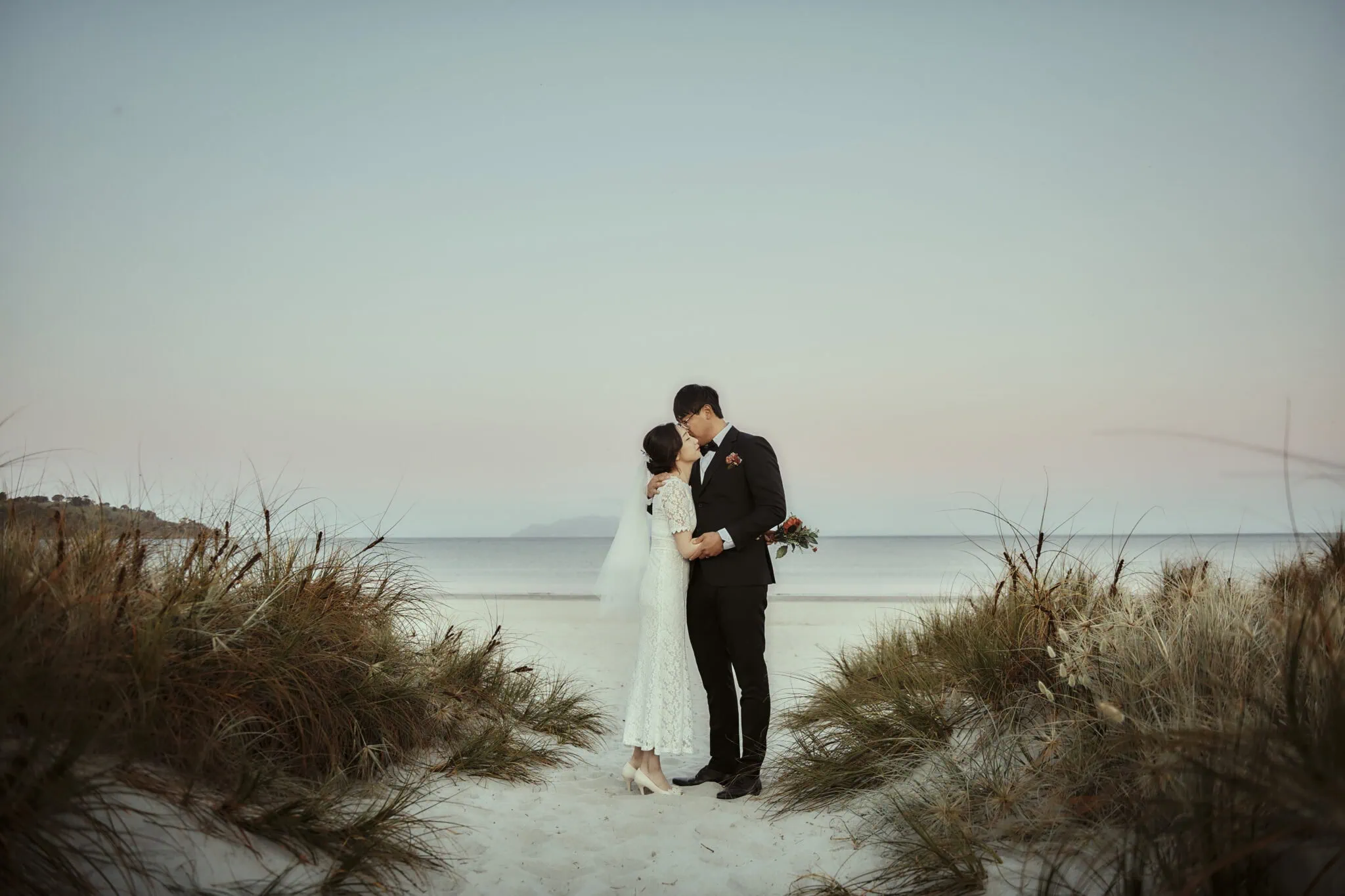 queenstown new zealand heli wedding elopement photographer videographer | Mijin and Joonsu, in elegant formal attire, embrace and kiss on a beach with grass-covered dunes in the foreground and the ocean in the background during sunset. This stunning pre-wedding shoot captures their love amid the serene beauty of NZ's coastline.