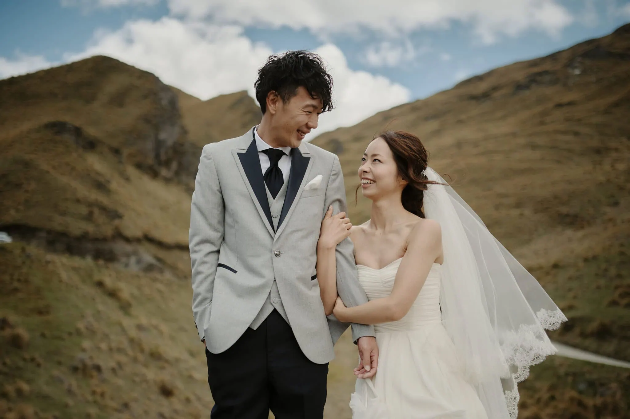 queenstown new zealand heli wedding elopement photographer videographer | A bride and groom stand outdoors in Queenstown, smiling at each other. The bride holds the groom's arm as they enjoy their picturesque Heli-Wedding. The background shows a rocky, hilly landscape under a partly cloudy sky.