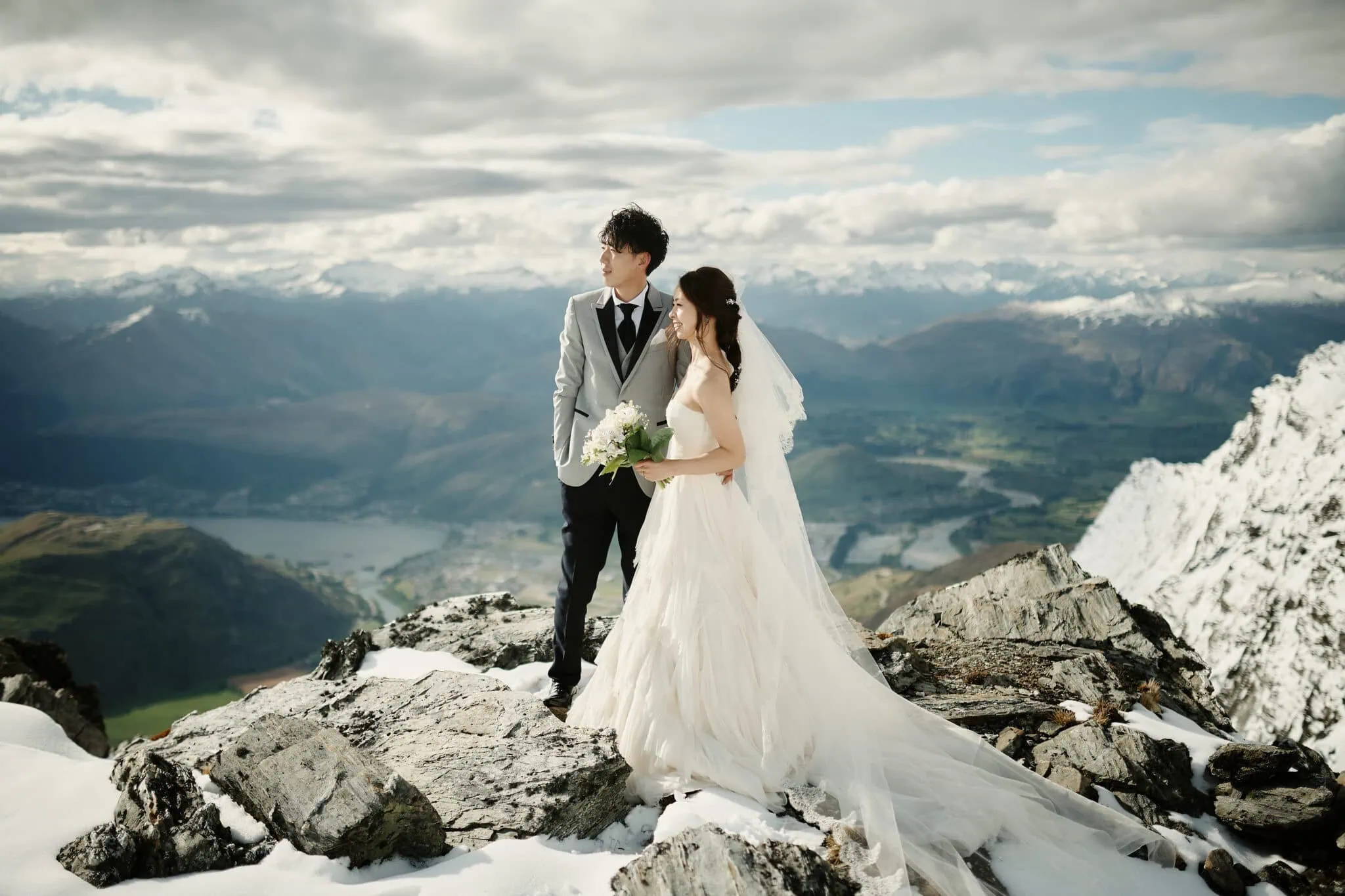 queenstown new zealand heli wedding elopement photographer videographer | Bride and groom standing on a rocky outcrop surrounded by snow-covered mountains near Queenstown, with a scenic valley and lake in the background. They are looking into the distance under a partly cloudy sky during their heli-wedding.