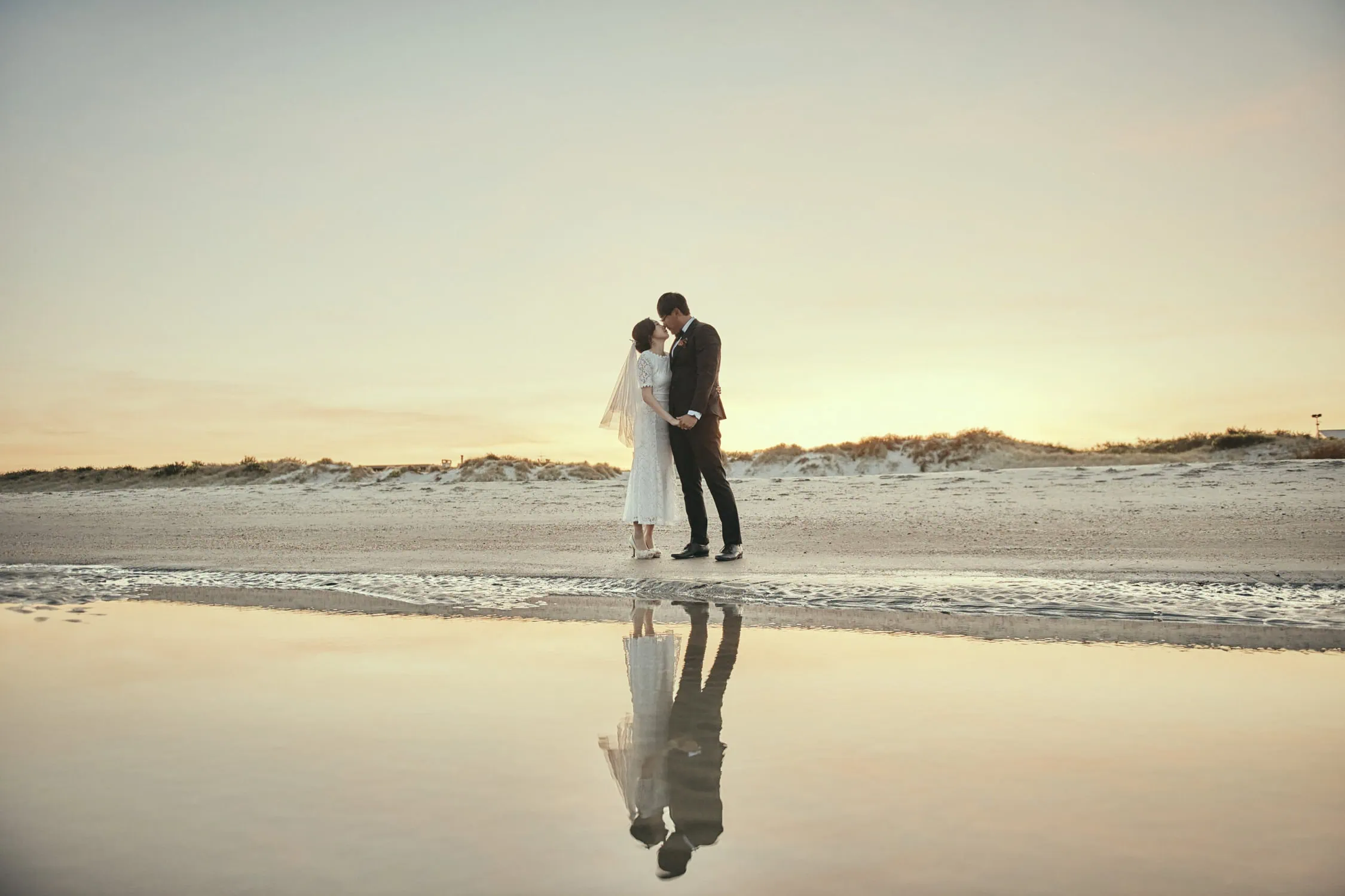 queenstown new zealand heli wedding elopement photographer videographer | Mijin and Joonsu stand on a beach at sunset, holding hands and kissing, with their reflection visible in a shallow pool of water—a perfect moment captured during their NZ pre-wedding shoot.