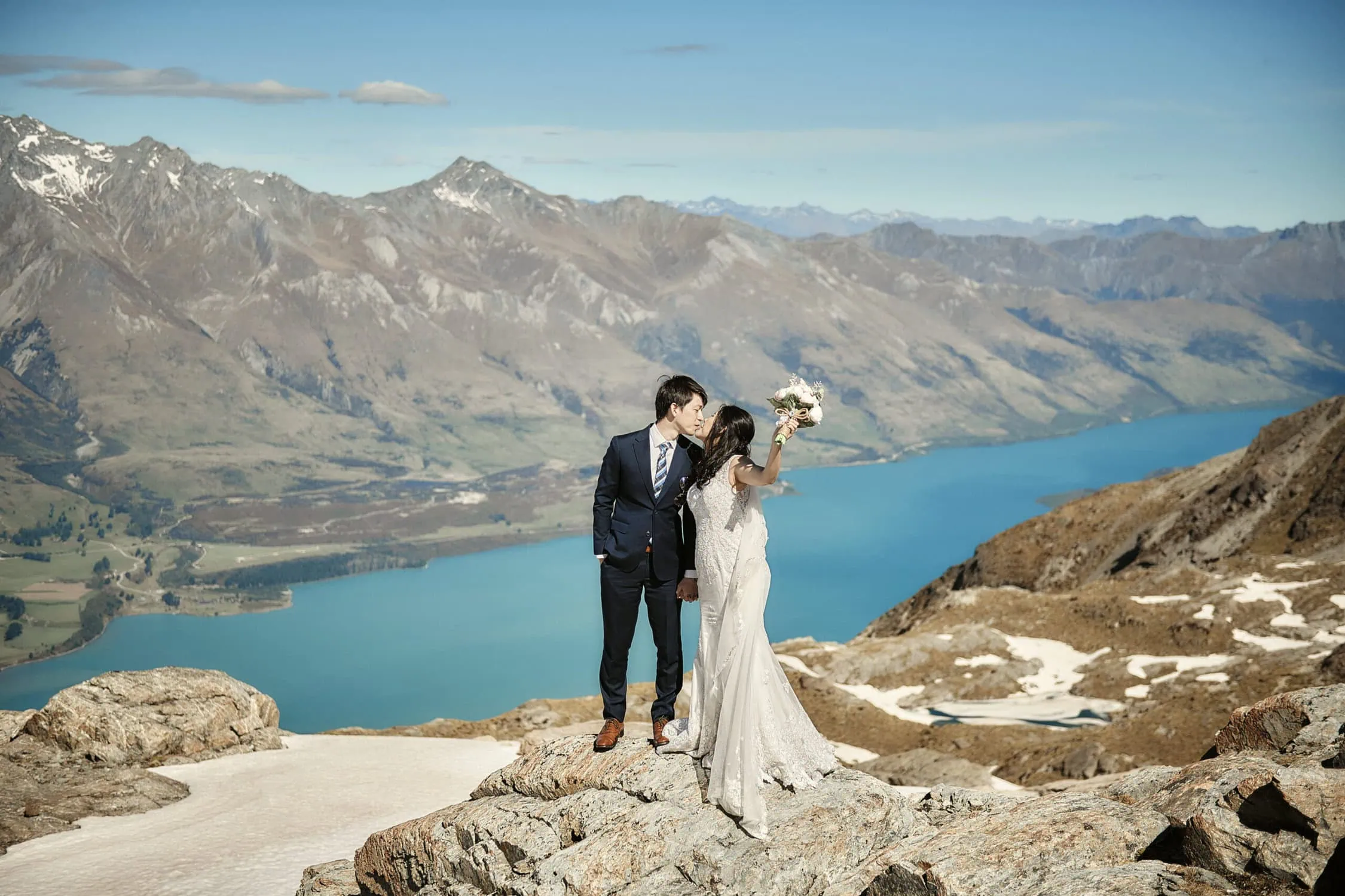 queenstown new zealand heli wedding elopement photographer videographer | A bride and groom stand on a rocky ledge with a breathtaking mountainous backdrop and the blue lake of Queenstown, New Zealand below, captured in a kiss during their pre-wedding shoot.