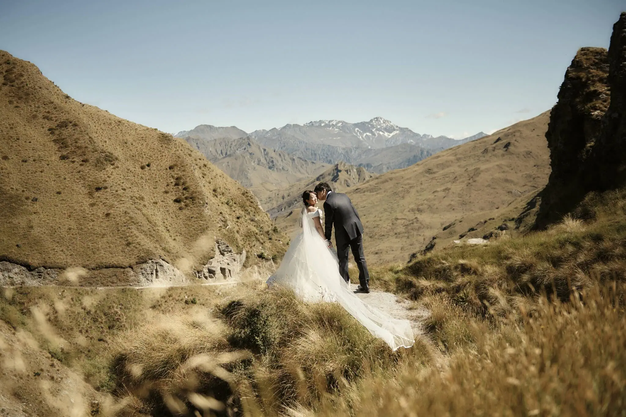 queenstown new zealand heli wedding elopement photographer videographer | A bride and groom share a kiss on a rocky hilltop with a mountainous landscape in the background, capturing the romance of their pre-wedding shoot near Tekapo.