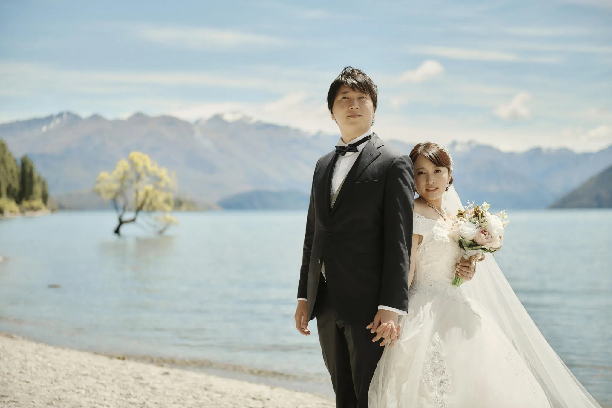 queenstown new zealand heli wedding elopement photographer videographer | A bride and groom stand hand-in-hand on a lake shore, with mountains in the background during their pre-wedding shoot. The bride holds a bouquet, and they both wear formal wedding attire, capturing the magic of Tekapo's serene beauty.