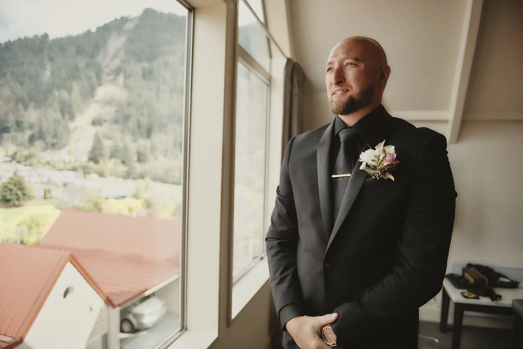 queenstown new zealand heli wedding elopement photographer videographer | A man in formal attire stands in front of a window with a view of Queenstown's mountainous landscape. He is wearing a black suit and a boutonniere, ready for an intimate elopement.