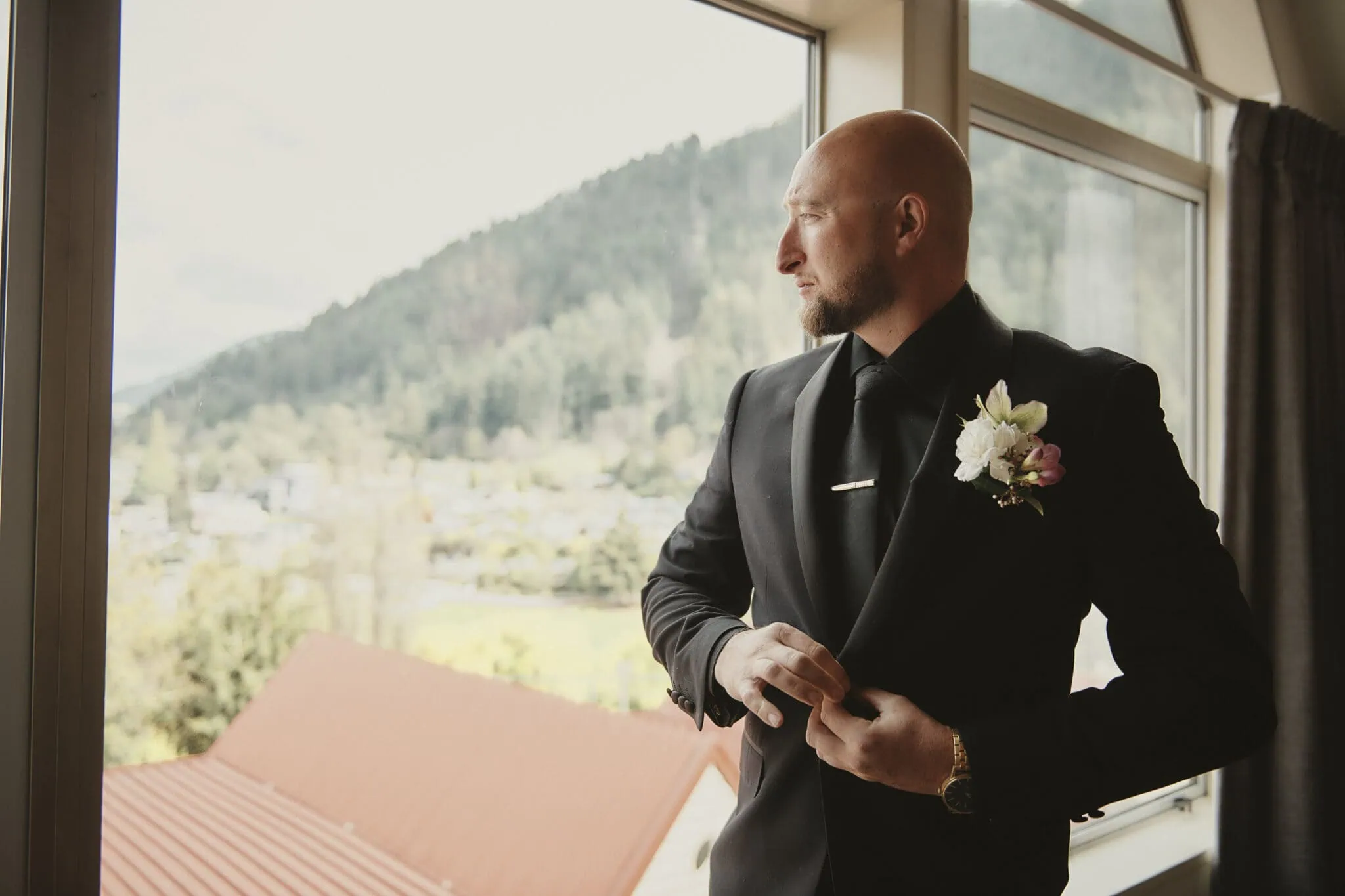 queenstown new zealand heli wedding elopement photographer videographer | A bald man in a black suit with a flower boutonniere stands by a window, adjusting his jacket, and looks outside. The scenic view of mountains and rooftops hints at a dreamy Queenstown elopement.