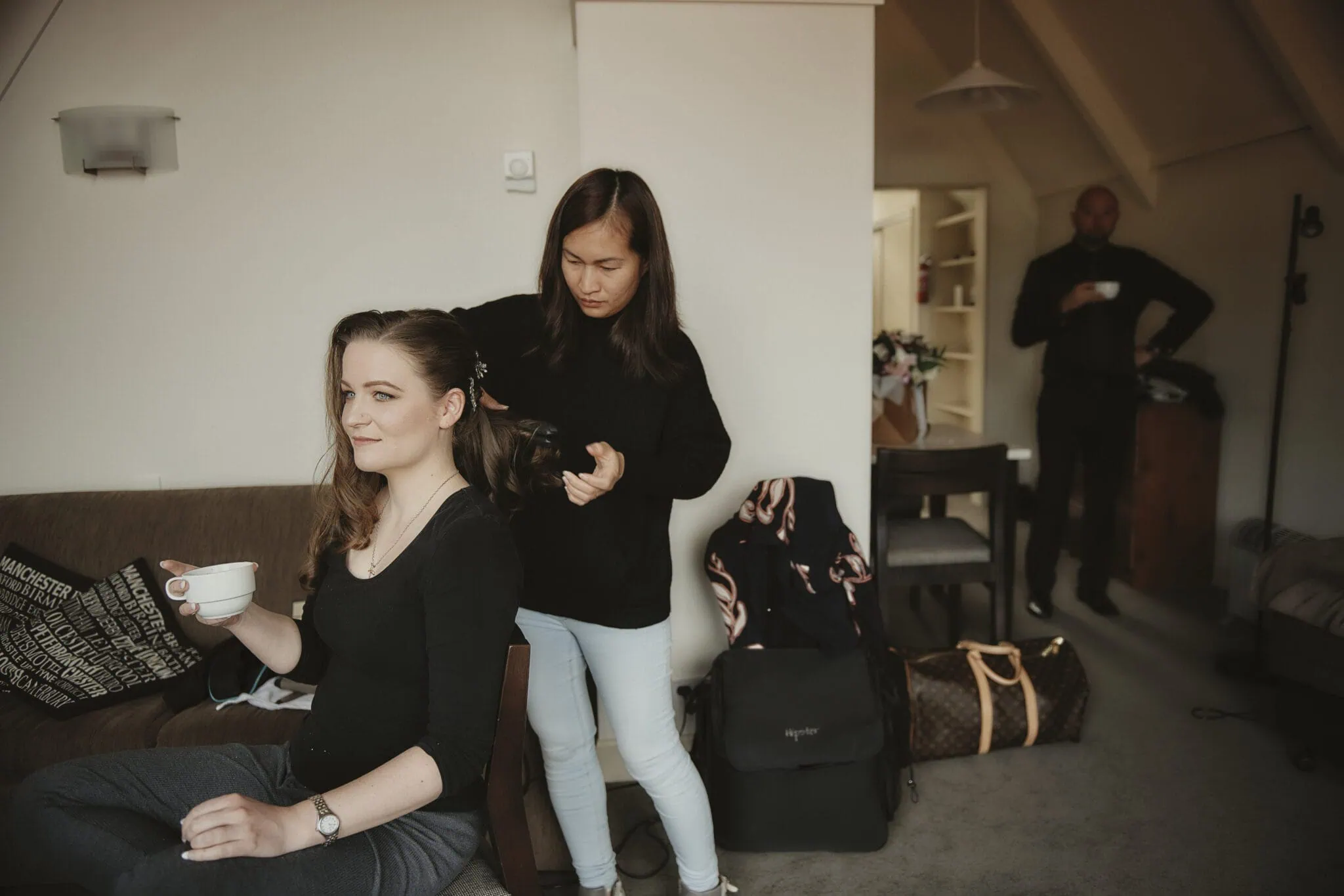 queenstown new zealand heli wedding elopement photographer videographer | A woman sits on a chair having her hair styled by another woman standing behind her, preparing for an elopement. She holds a cup in her hand in a room with bags and furniture in the background. Another person stands nearby, ready to assist with their Queenstown heli-wedding adventures.