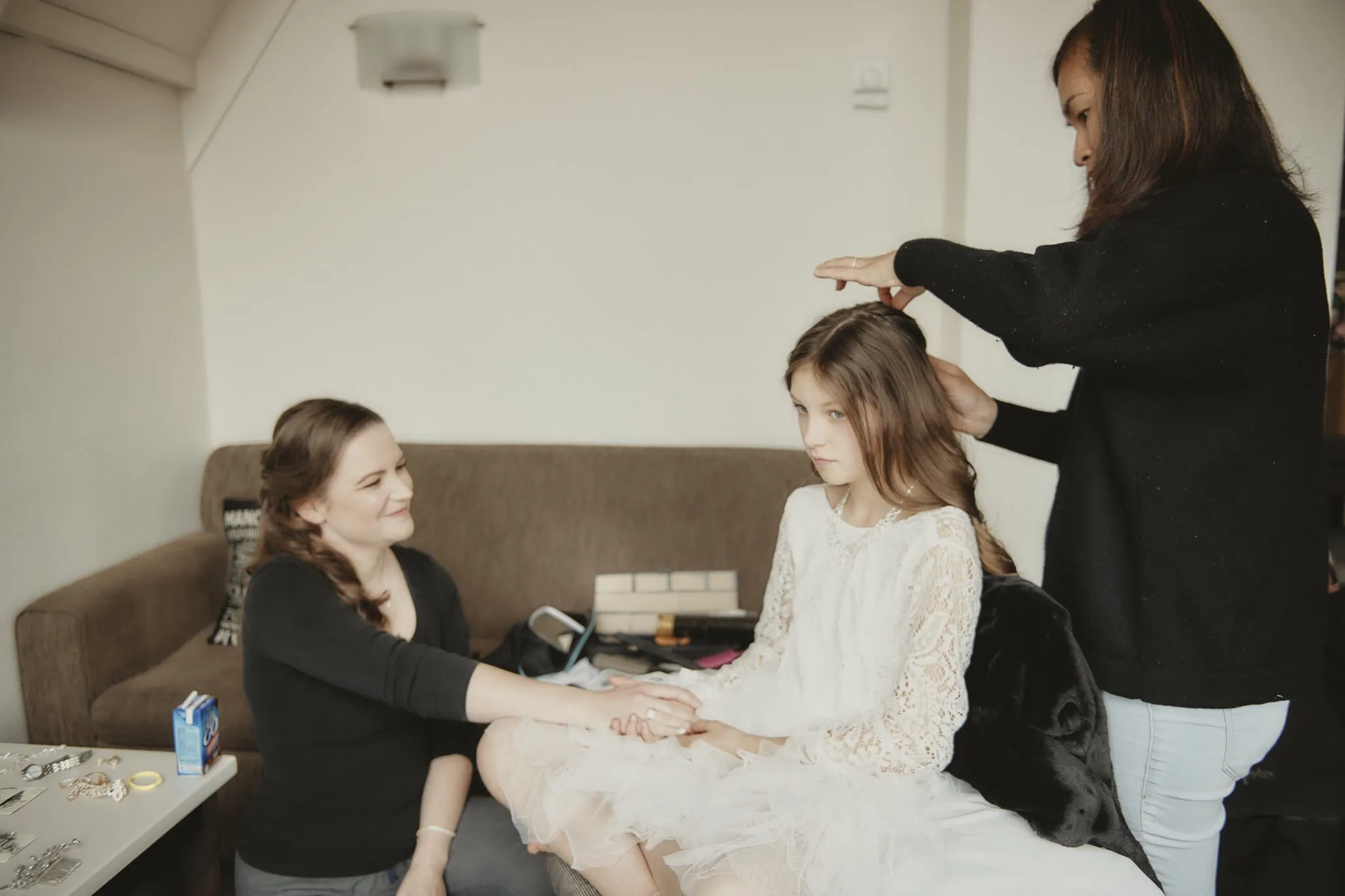 queenstown new zealand heli wedding elopement photographer videographer | Two women style a young girl's hair with the care and precision you might expect at a Queenstown heli-wedding. The girl, seated in a white dress, looks straight ahead while they work. One woman leans forward, smiling, and holding the girl's hand.
