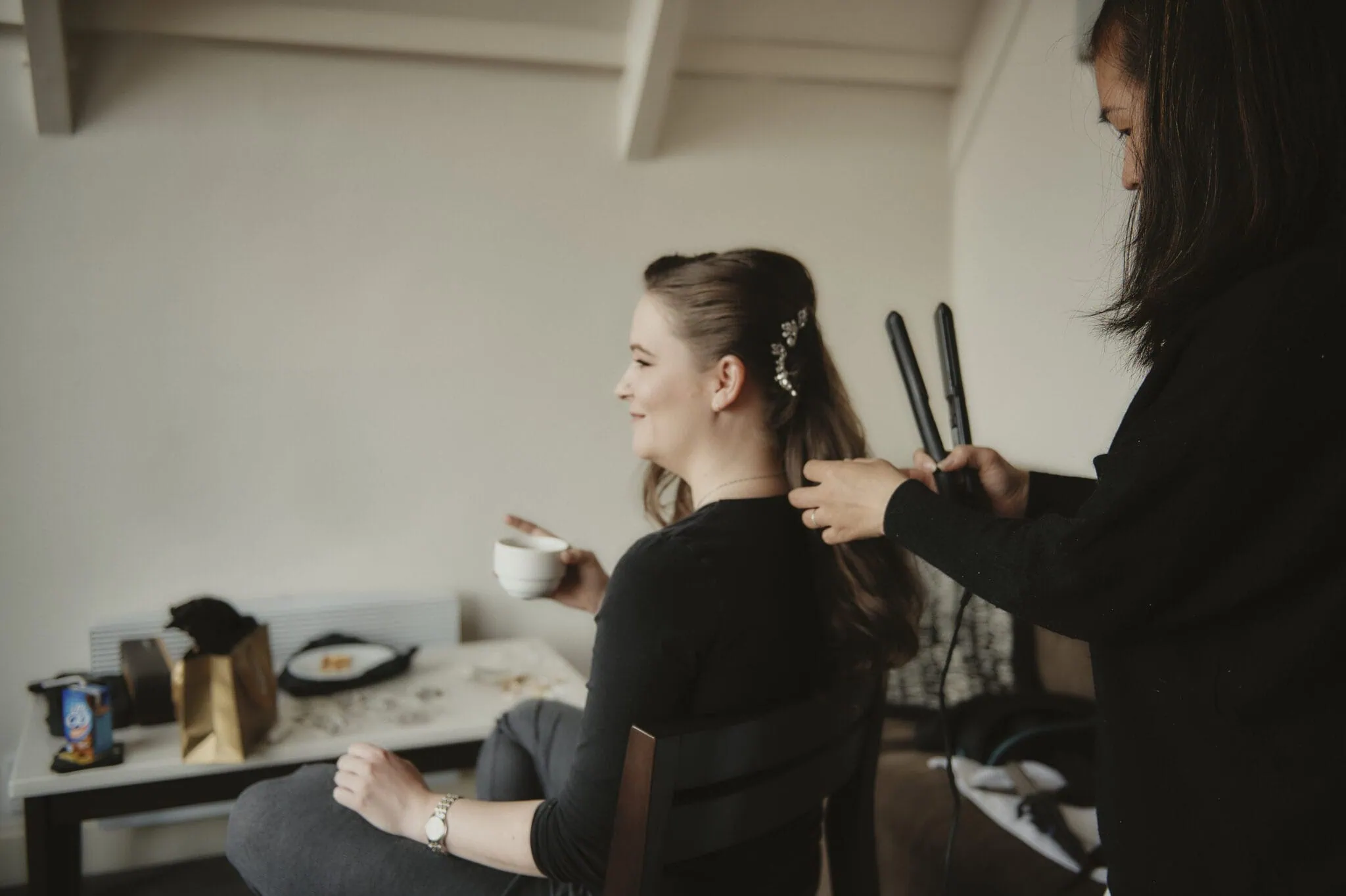 queenstown new zealand heli wedding elopement photographer videographer | A woman sits in a chair holding a cup while another woman styles her hair with a flat iron, preparing her for a magical Heli-Wedding in Queenstown.
