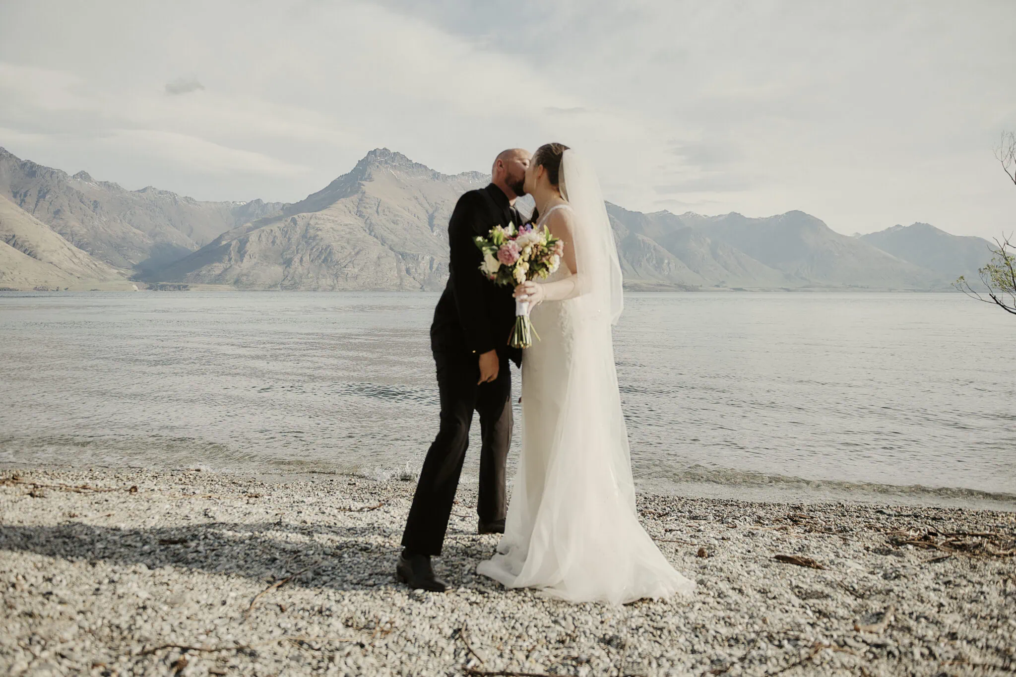 queenstown new zealand heli wedding elopement photographer videographer | A newlywed couple, the groom in black and the bride in a white dress with a veil, kiss on a pebble beach in Queenstown with a lake and mountains as their backdrop. The bride holds a bouquet, capturing the romance of their intimate elopement.