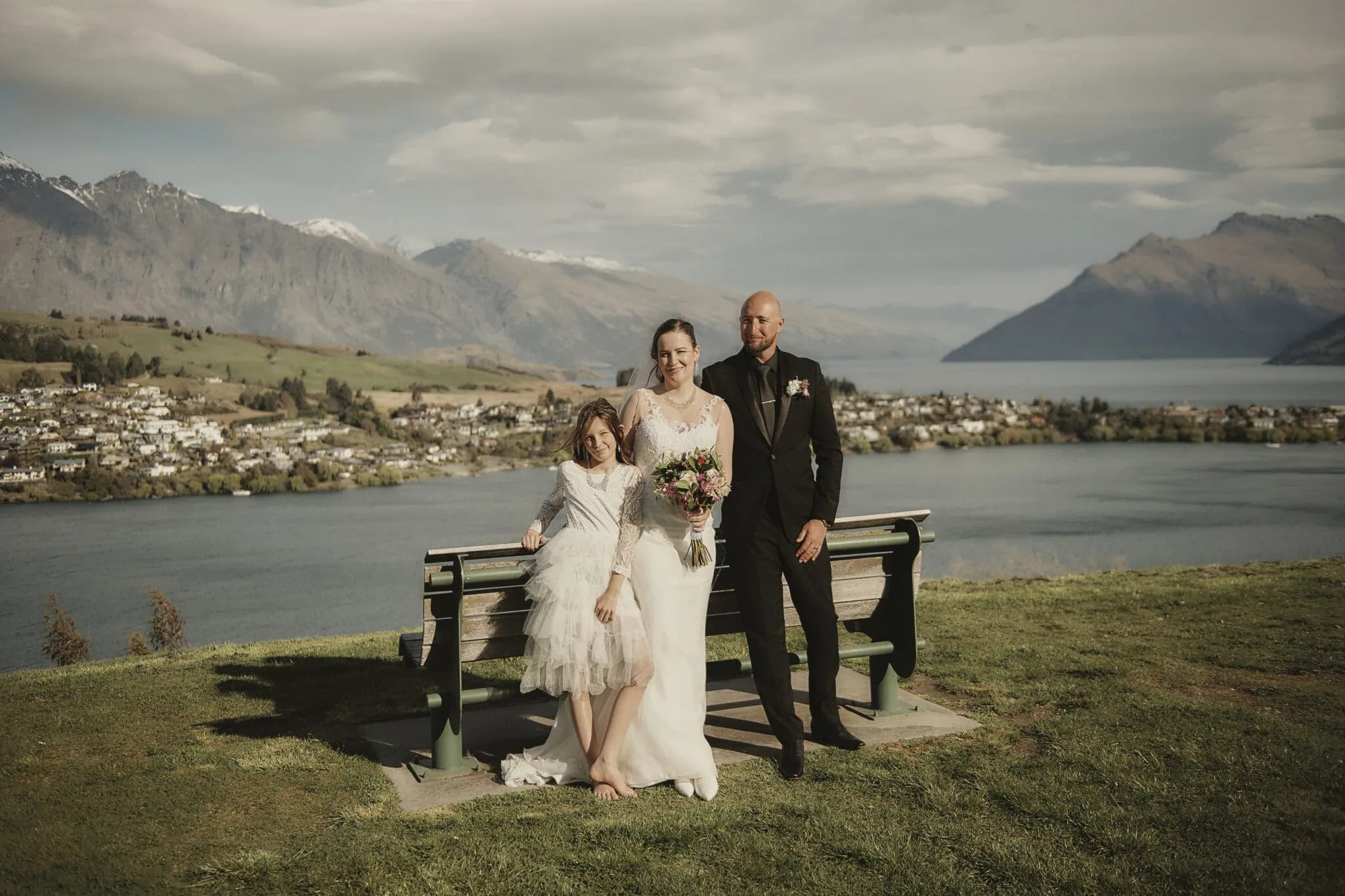 queenstown new zealand heli wedding elopement photographer videographer | A woman in a white dress holds a bouquet, standing with a man in a suit and a child in a dress, framed by the scenic beauty of Queenstown's lake and mountains. They stand by a bench on the grassy area, embodying the serene charm of an intimate elopement.