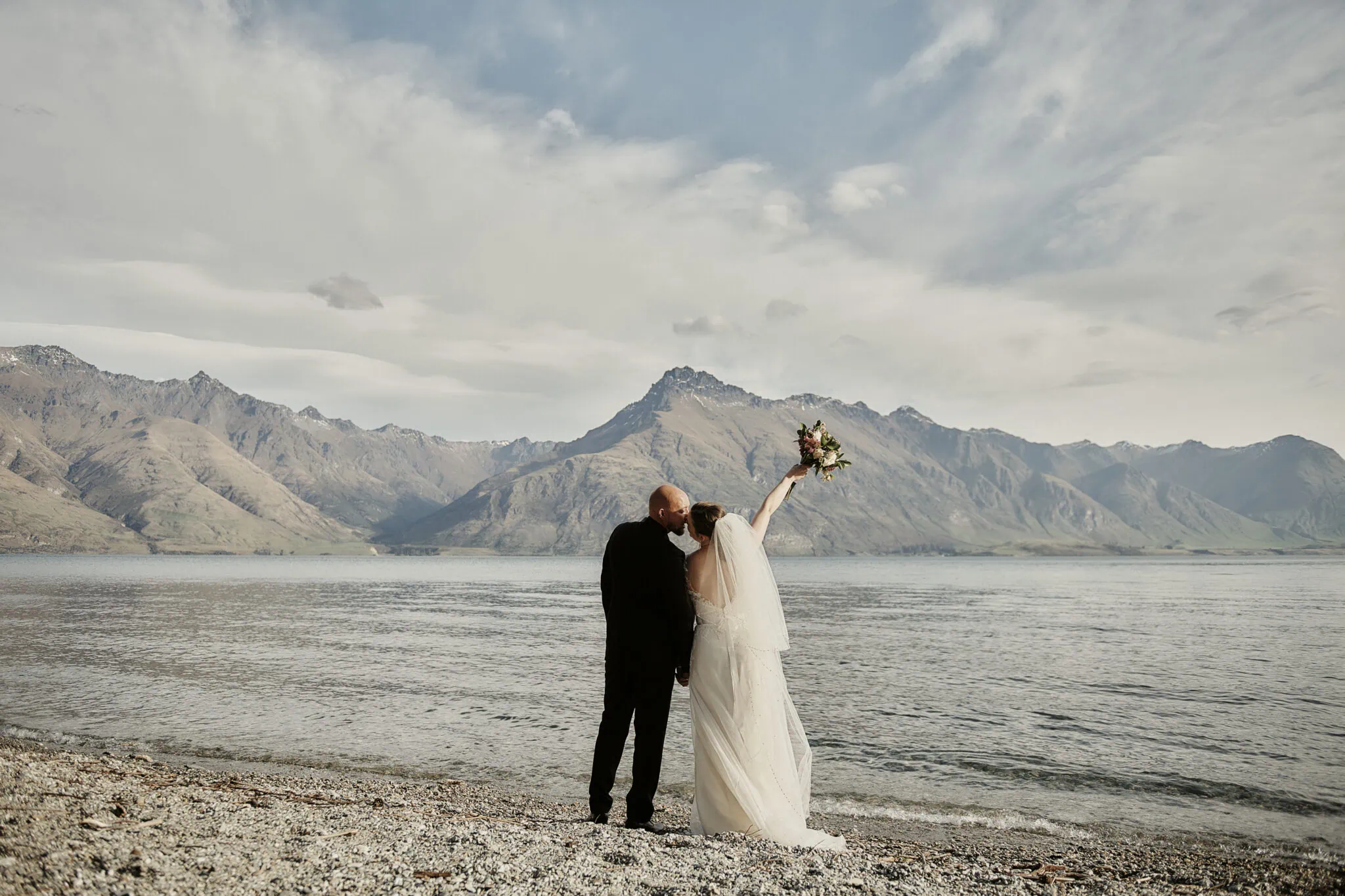 queenstown new zealand heli wedding elopement photographer videographer | A couple in wedding attire stands on a pebble beach by a lake in Queenstown, with mountains in the background. The bride raises her bouquet into the air, celebrating their adventurous elopement.