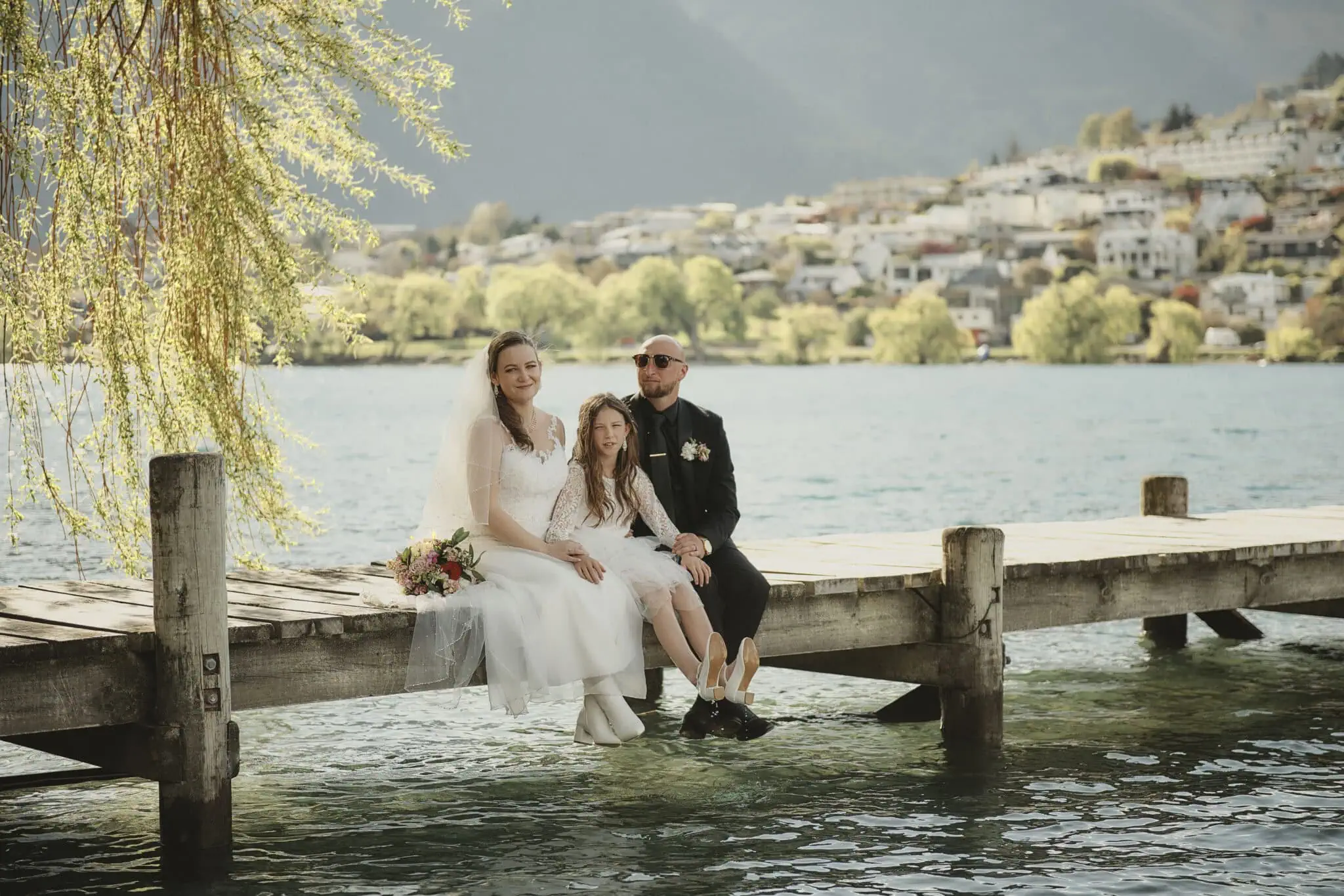 queenstown new zealand heli wedding elopement photographer videographer | A bride and groom sit on a dock with a young girl, likely their daughter, by a lake with Queenstown's scenic town and mountains in the background.
