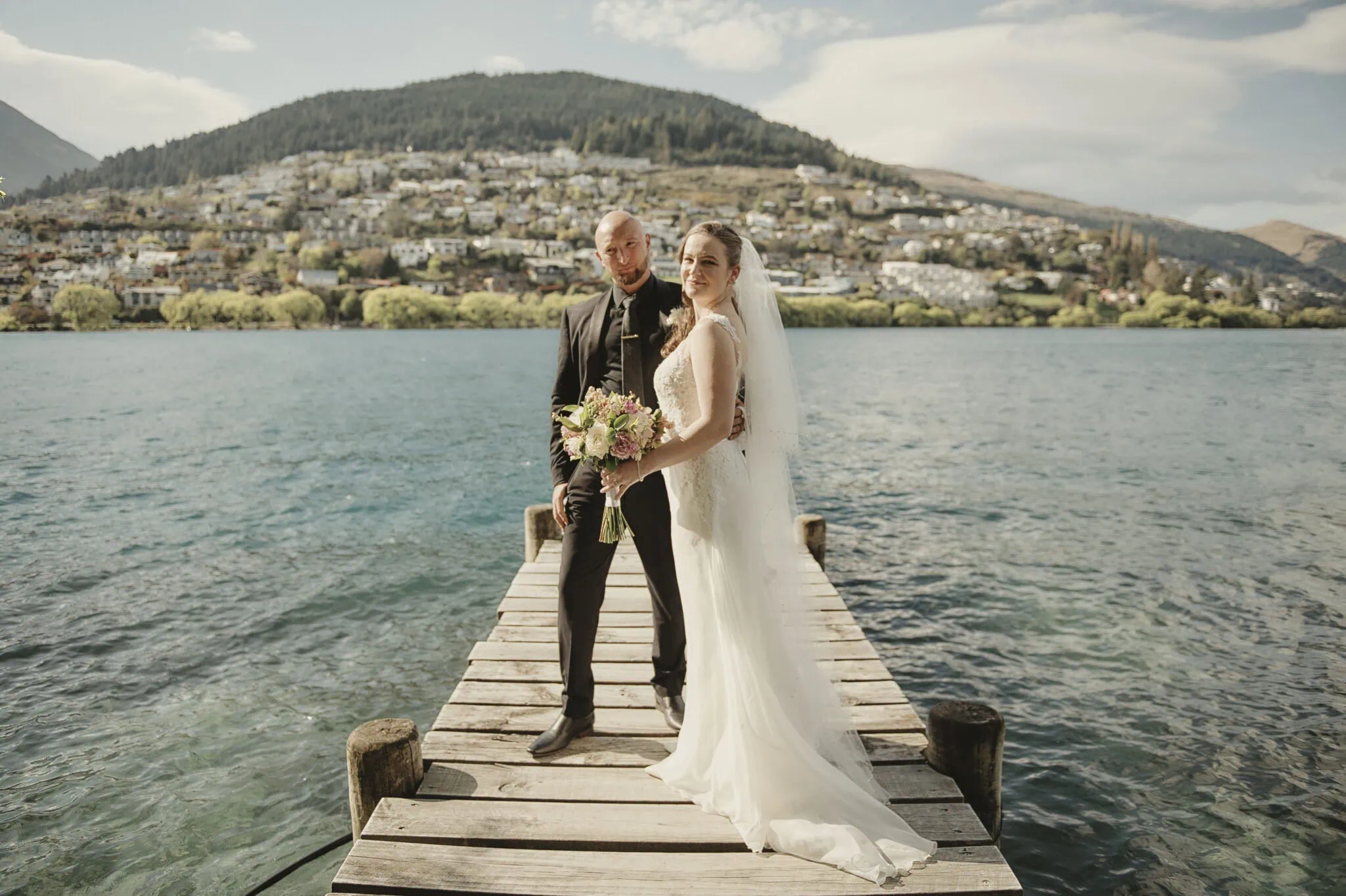 queenstown new zealand heli wedding elopement photographer videographer | A bride and groom stand on a wooden dock by a lake with a mountainous landscape and the charming town of Queenstown in the background. The bride is holding a bouquet and wearing a white dress, while the groom wears a black suit, capturing the magic of an NZ heli-wedding.