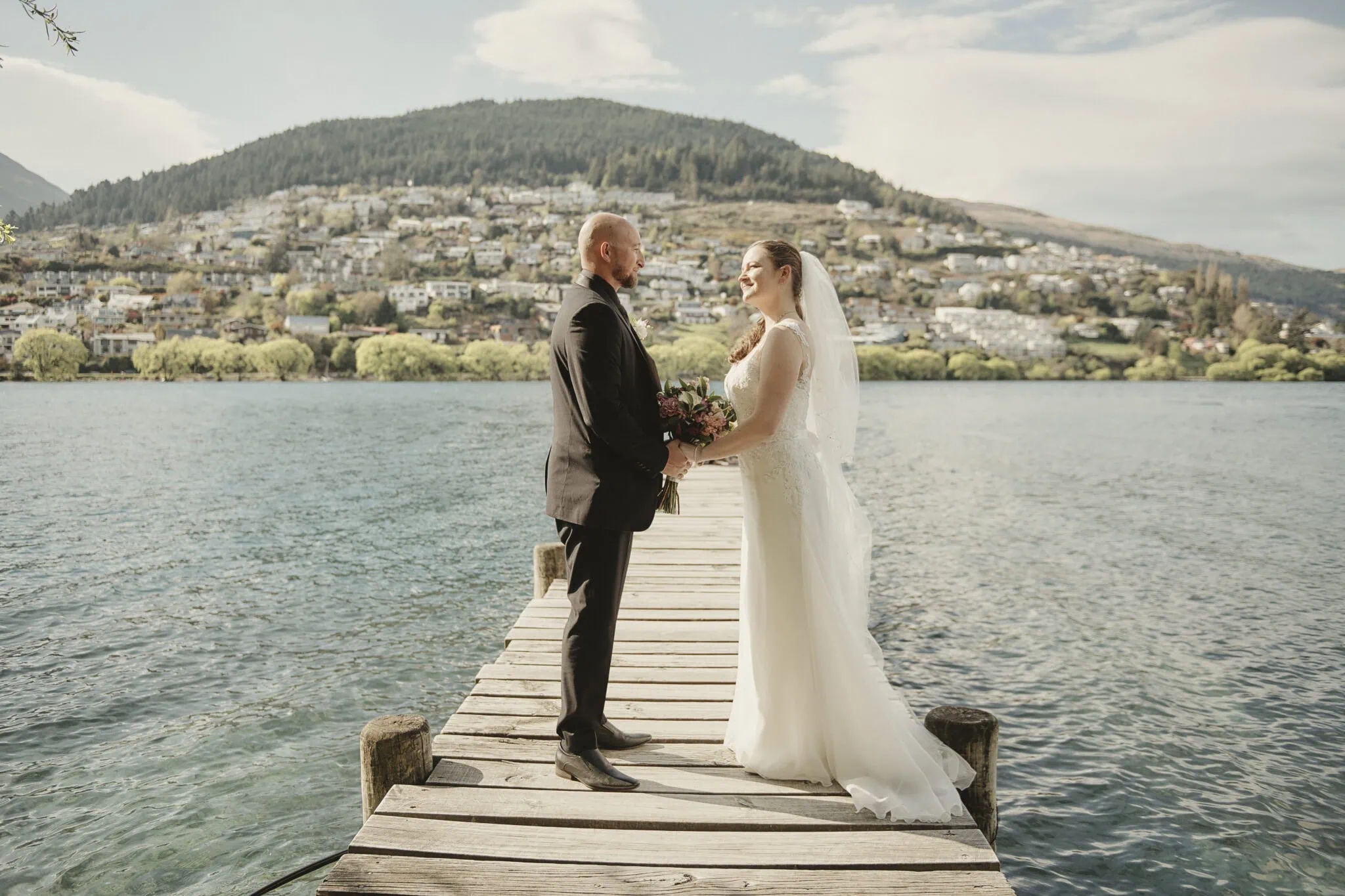 queenstown new zealand heli wedding elopement photographer videographer | A bride and groom stand facing each other on a wooden dock by a lake in stunning Queenstown, with a hilly landscape and scattered houses in the background, capturing the intimate essence of an elopement.
