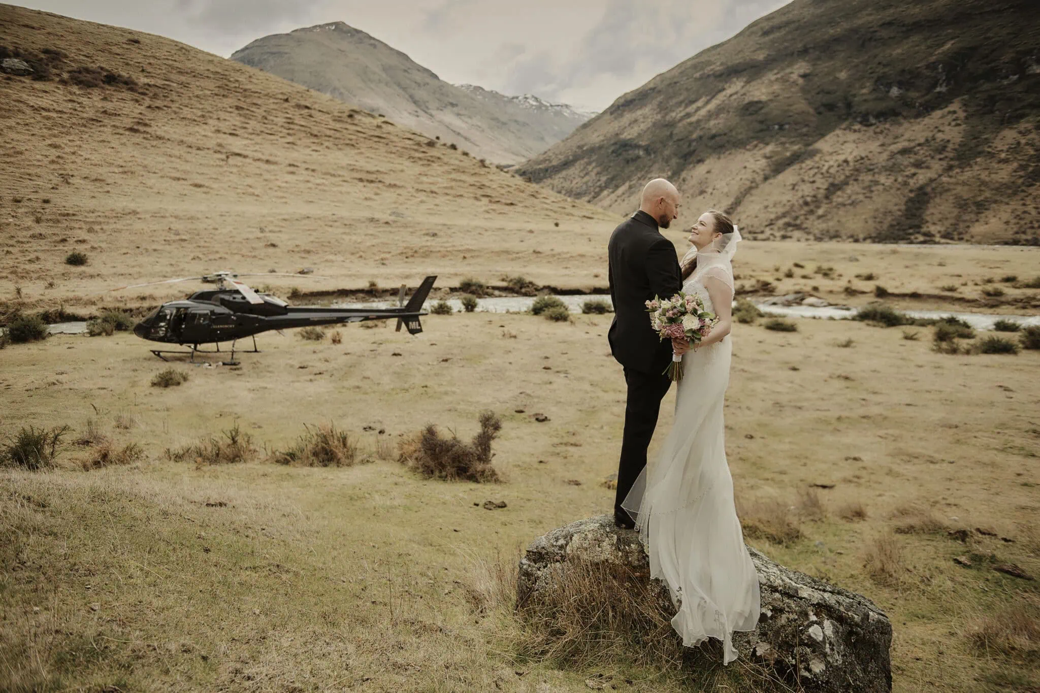 queenstown new zealand heli wedding elopement photographer videographer | A bride and groom stand on a rock in the stunning Queenstown mountainous landscape, with a helicopter hinting at their adventurous Heli-Wedding in the background. The bride holds a bouquet, and both are dressed elegantly for their unforgettable elopement.