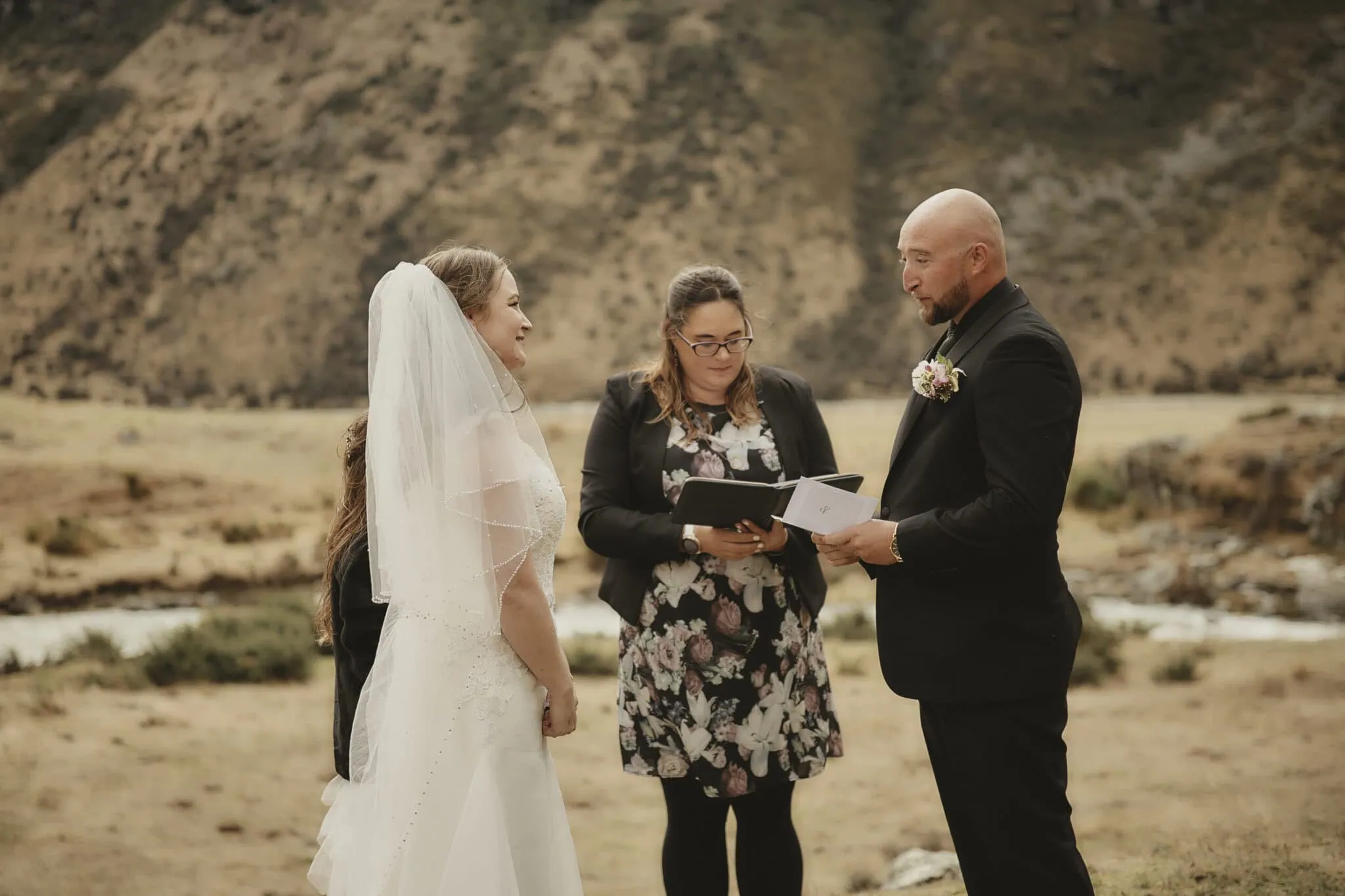 queenstown new zealand heli wedding elopement photographer videographer | A couple stands outdoors near a river and mountainous landscape in Queenstown, exchanging vows with an officiant present. The bride wears a veil, and the groom holds a booklet while the officiant reads. This picturesque setting is perfect for their NZ heli-wedding, adding an unforgettable touch to their special day.