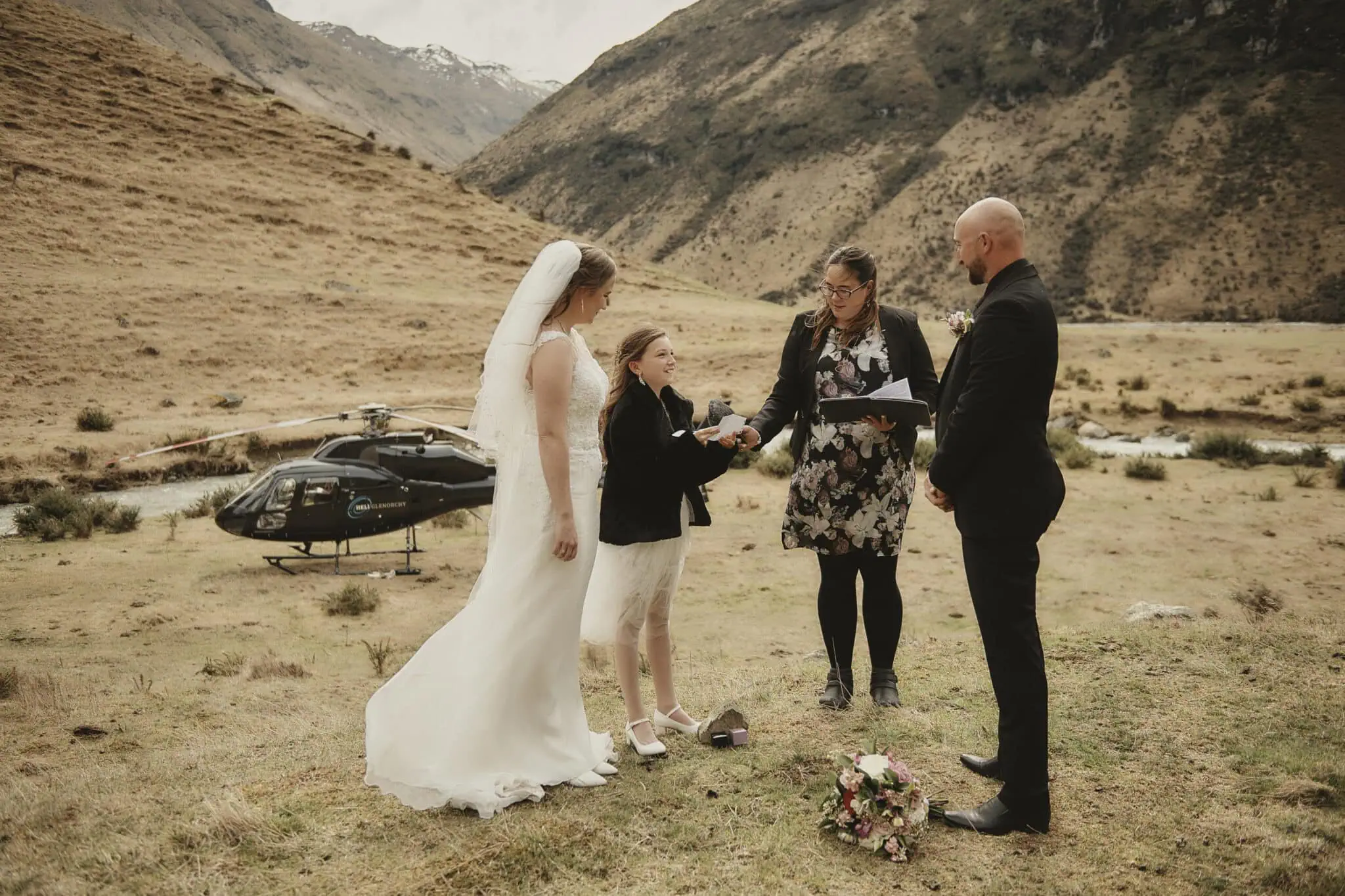 queenstown new zealand heli wedding elopement photographer videographer | A bride and groom stand with a young girl and an officiant in an outdoor elopement ceremony in Queenstown's mountainous area. A helicopter, hinting at their unique heli-wedding, is parked in the background. The bride holds a bouquet of flowers.