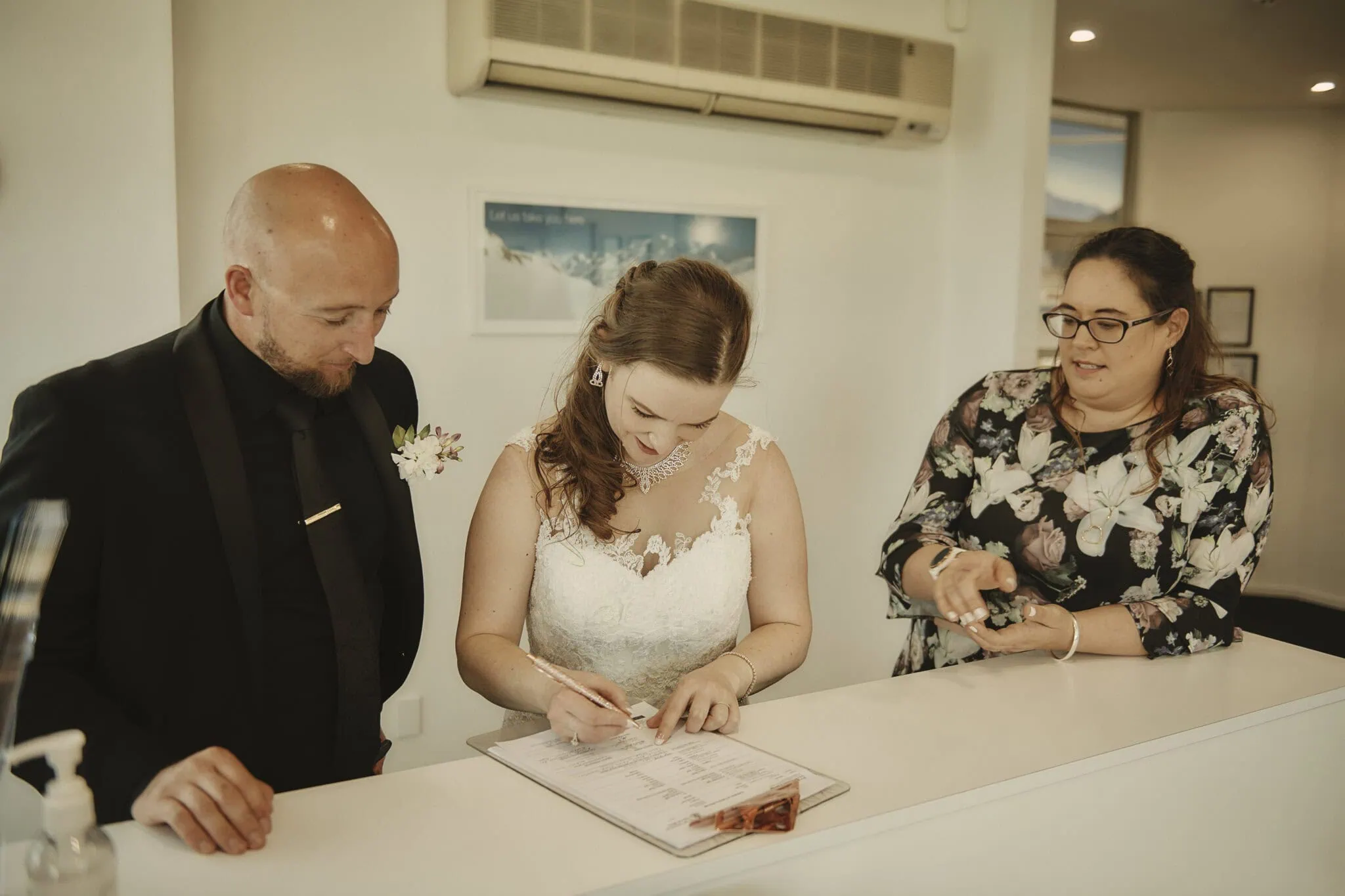 queenstown new zealand heli wedding elopement photographer videographer | A bride in a white lace dress signs a document at a counter, accompanied by a bald man in black attire and a woman in a floral dress, fresh from their unforgettable Heli-Wedding adventure in Queenstown, NZ.