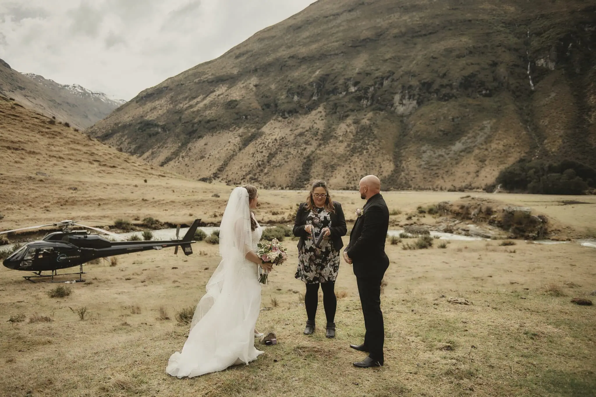 queenstown new zealand heli wedding elopement photographer videographer | A couple stands with an officiant during their outdoor elopement in a stunning mountainous landscape near Queenstown. A helicopter is parked nearby on the grass, completing the perfect heli-wedding scene.