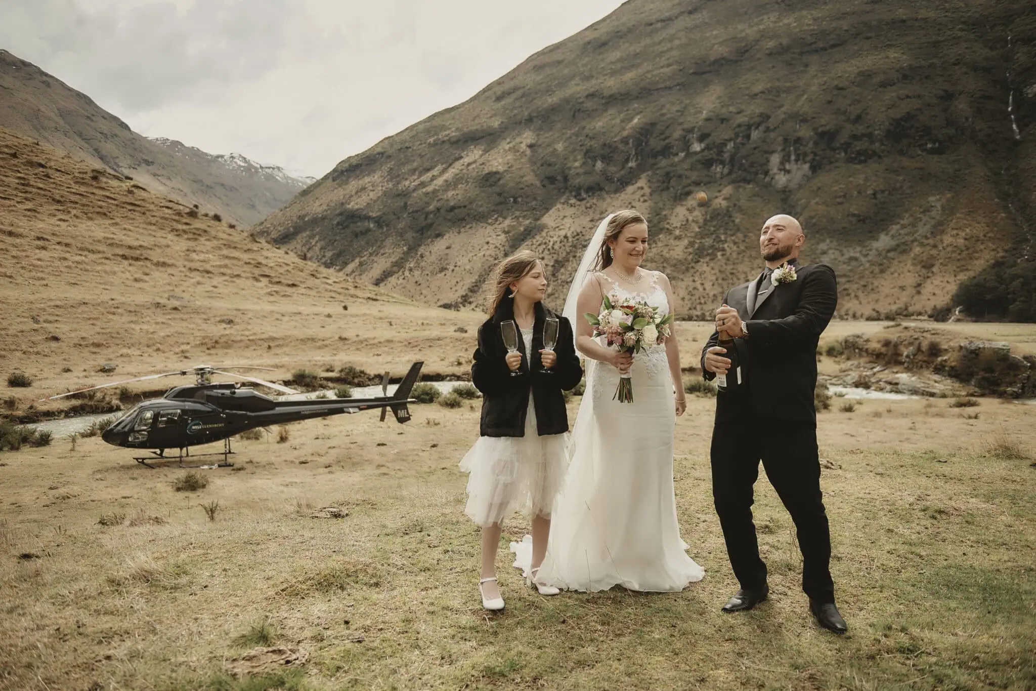 queenstown new zealand heli wedding elopement photographer videographer | A bride and groom, accompanied by a young girl, stand in a scenic mountainous area near Queenstown with a helicopter in the background. Celebrating their elopement, the man is opening a bottle of champagne while the others watch. This heli-wedding captures an unforgettable moment against nature's breathtaking backdrop.