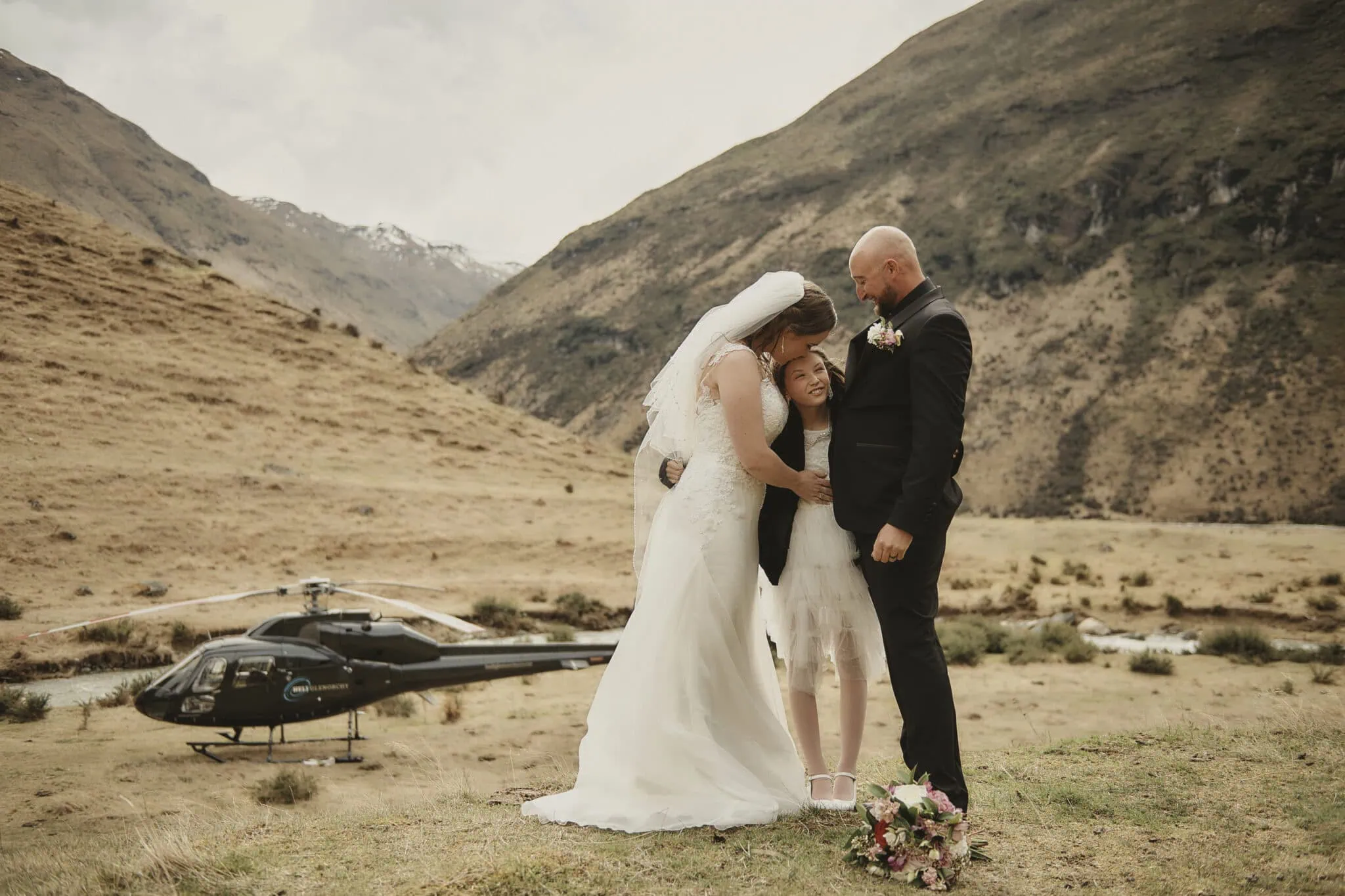queenstown new zealand heli wedding elopement photographer videographer | A bride and groom stand with a child in an outdoor mountainous landscape in Queenstown. A bouquet lies on the ground, and a helicopter is parked nearby, capturing the essence of their heli-wedding elopement.