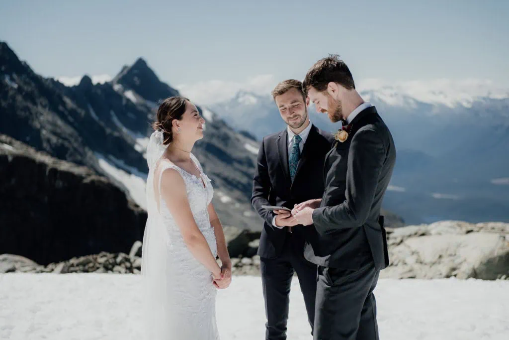 queenstown new zealand heli wedding elopement photographer videographer | A couple, dressed in wedding attire, stands on a snowy mountain with a man officiating their ceremony. Snow-covered peaks rise in the background under a clear sky, capturing the magic of an Earnslaw Burn heli-wedding near Queenstown.