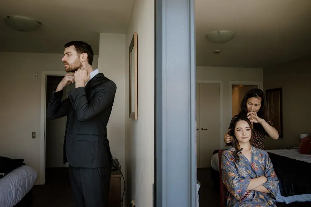 queenstown new zealand heli wedding elopement photographer videographer | A man adjusts his collar in a bedroom while a woman sits in a chair getting her hair styled by another woman, all preparing for their stunning Heli-Wedding at Earnslaw Burn, Queenstown NZ.