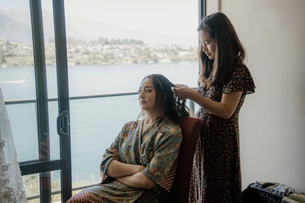 queenstown new zealand heli wedding elopement photographer videographer | A woman stands and styles the hair of another woman who is seated with arms crossed. They are next to a window overlooking the breathtaking Earnslaw Burn in Queenstown, creating a serene backdrop for their intimate heli-wedding prep.