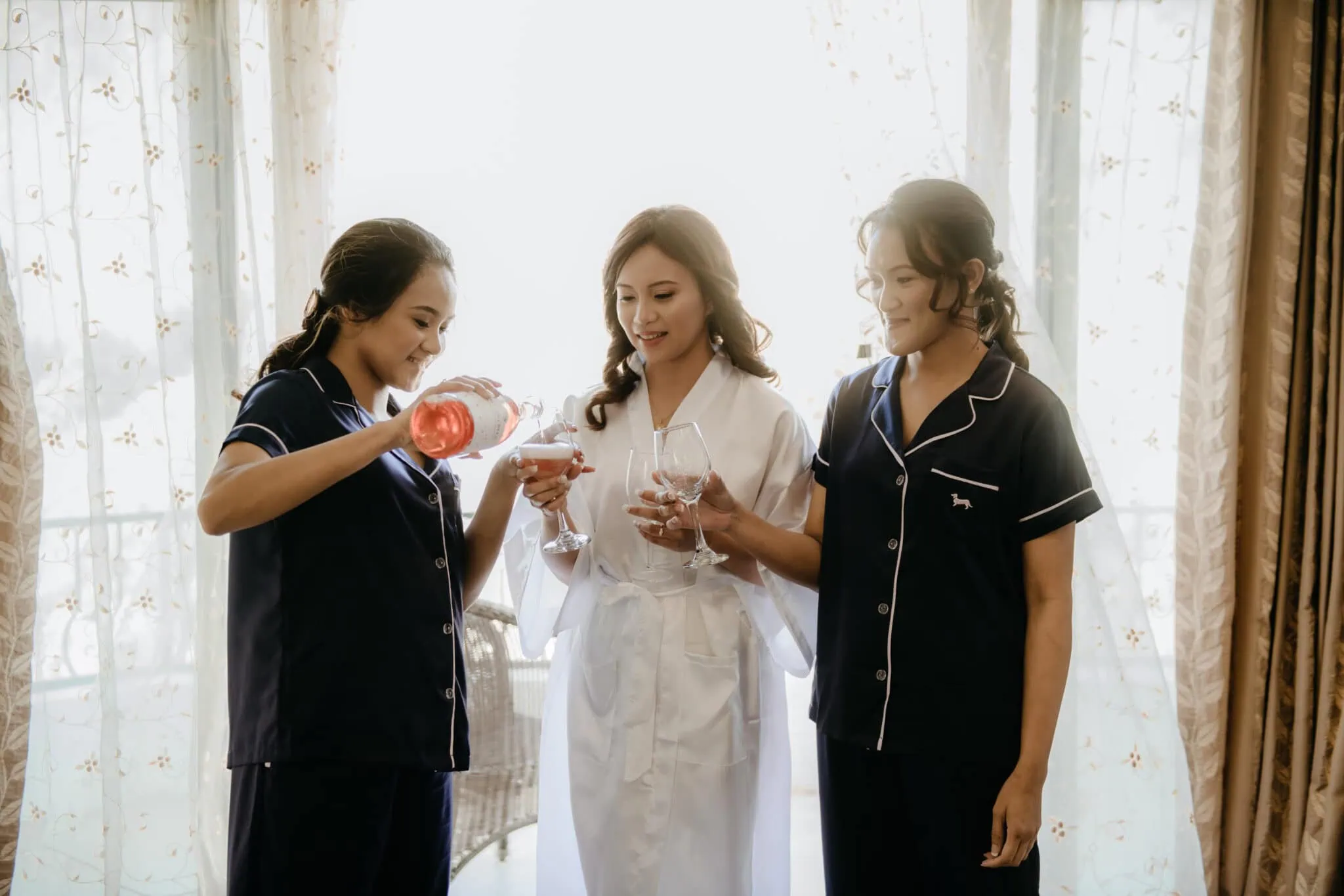 queenstown new zealand heli wedding elopement photographer videographer | Three women in pajamas stand together; one is pouring wine into two glasses. The central woman, in a white robe, holds a glass and smiles. They are in a room with curtains in the background, preparing for the Stoneridge Wedding of Natalya & John.
