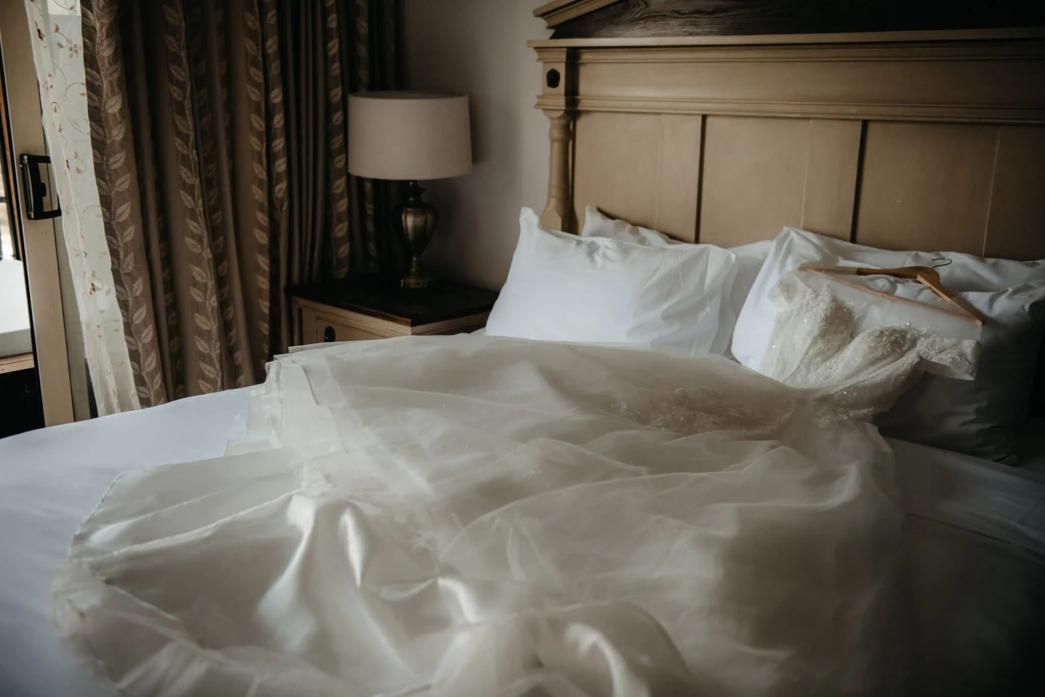 queenstown new zealand heli wedding elopement photographer videographer | A white wedding dress with lace details is laid out on a neatly made bed in a room at Stoneridge Wedding in Queenstown, NZ, featuring a beige headboard, nightstand, lamp, and curtains.