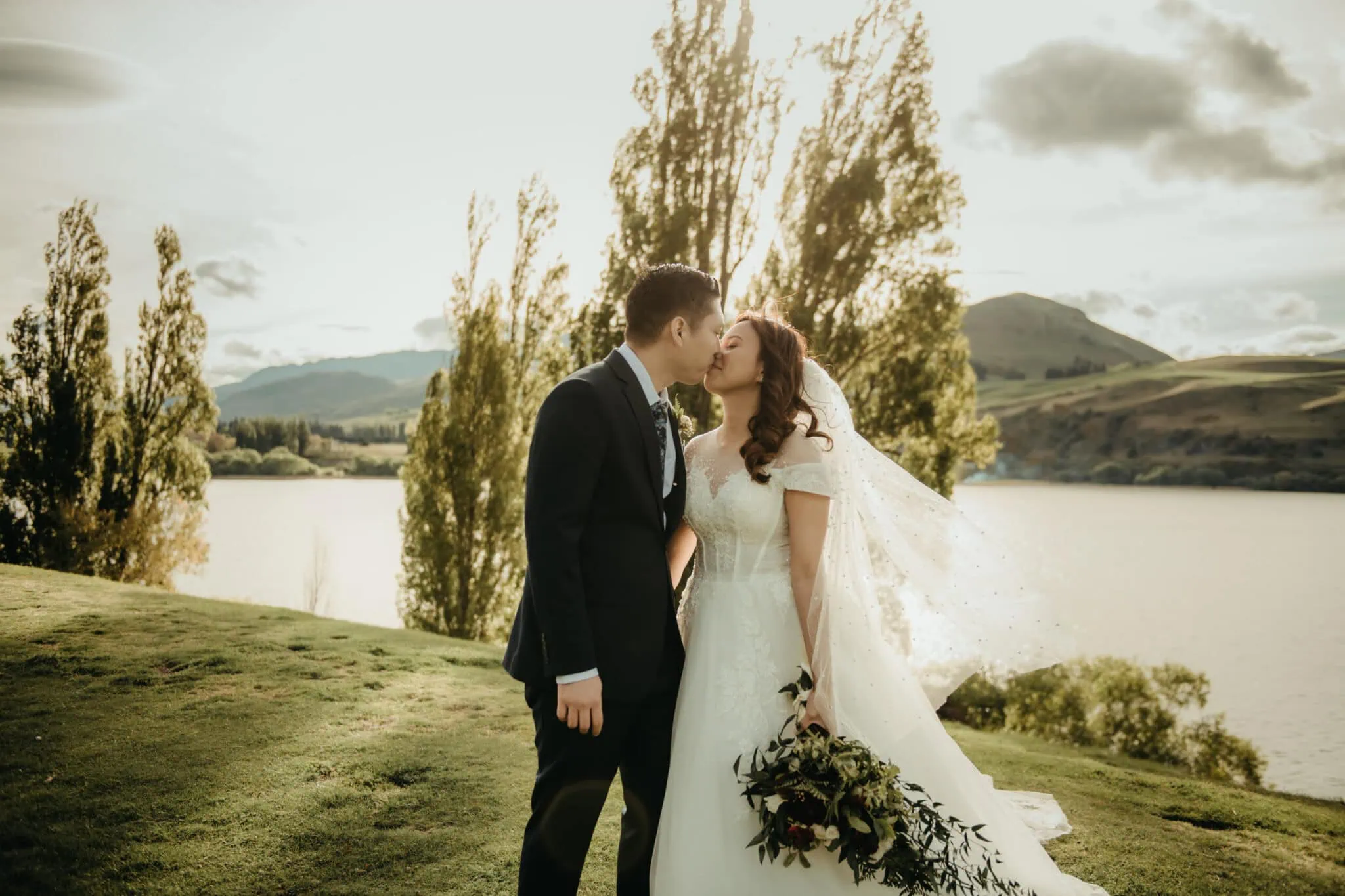 queenstown new zealand heli wedding elopement photographer videographer | A couple in wedding attire stands outdoors near a lake in Queenstown, kissing. Natalya holds a bouquet and wears a veil, while John dons a dark suit. Trees and hills form the beautiful backdrop of their Stoneridge Wedding.