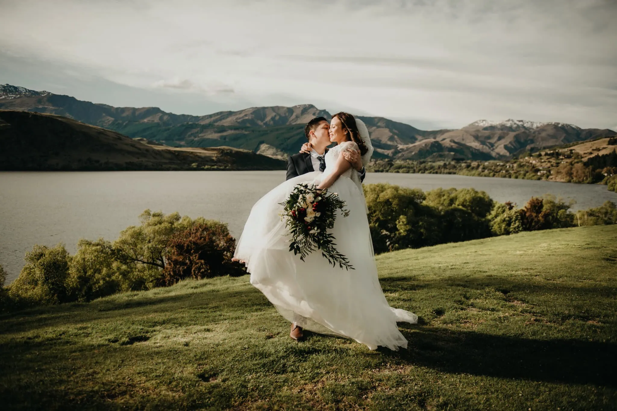 queenstown new zealand heli wedding elopement photographer videographer | A groom lifts his bride as they kiss in the picturesque setting of Stoneridge Wedding in Queenstown, with a serene lake, rolling hills, and a cloudy sky in the background. Natalya & John share a moment of pure joy while the bride holds a bouquet of flowers.
