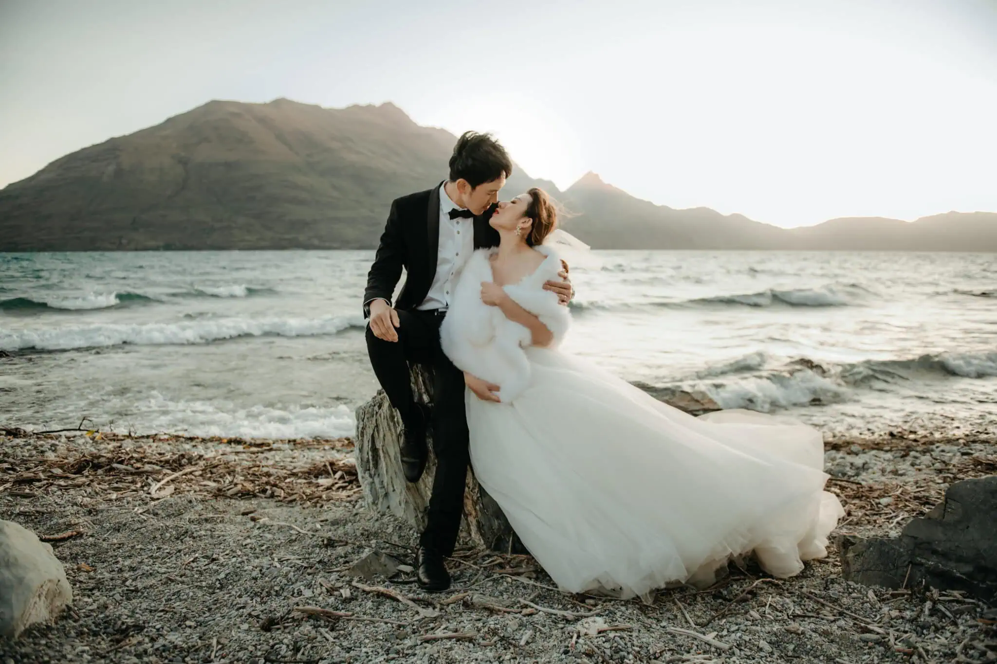 queenstown new zealand heli wedding elopement photographer videographer | A bride in a white wedding dress sits on a driftwood stump beside a groom in a black suit on a rocky beach, with mountains and a body of water in the background at sunset. This stunning pre-wedding shoot near Queenstown captures the essence of romance amid nature's splendor.