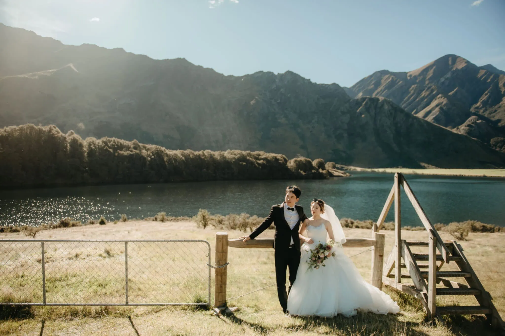 queenstown new zealand heli wedding elopement photographer videographer | Bride and groom standing by a wooden fence near a lake with a mountainous landscape in the background, capturing their serene Heli Pre-Wedding moment in Queenstown. The groom is in a black suit, and the bride is in a white gown holding a bouquet.