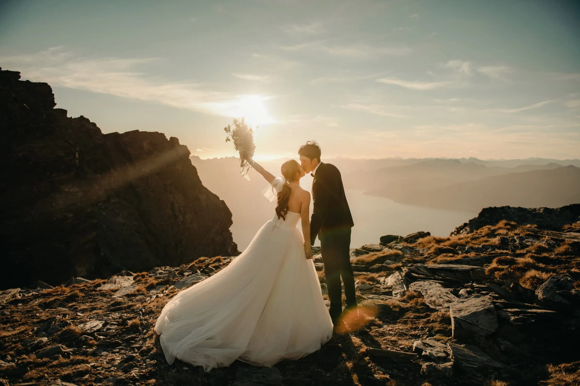 queenstown new zealand heli wedding elopement photographer videographer | A couple in wedding attire shares a kiss on a rocky mountaintop at sunset during their heli pre-wedding shoot in Queenstown, NZ. The bride holds a bouquet, and the landscape below is illuminated by the setting sun.