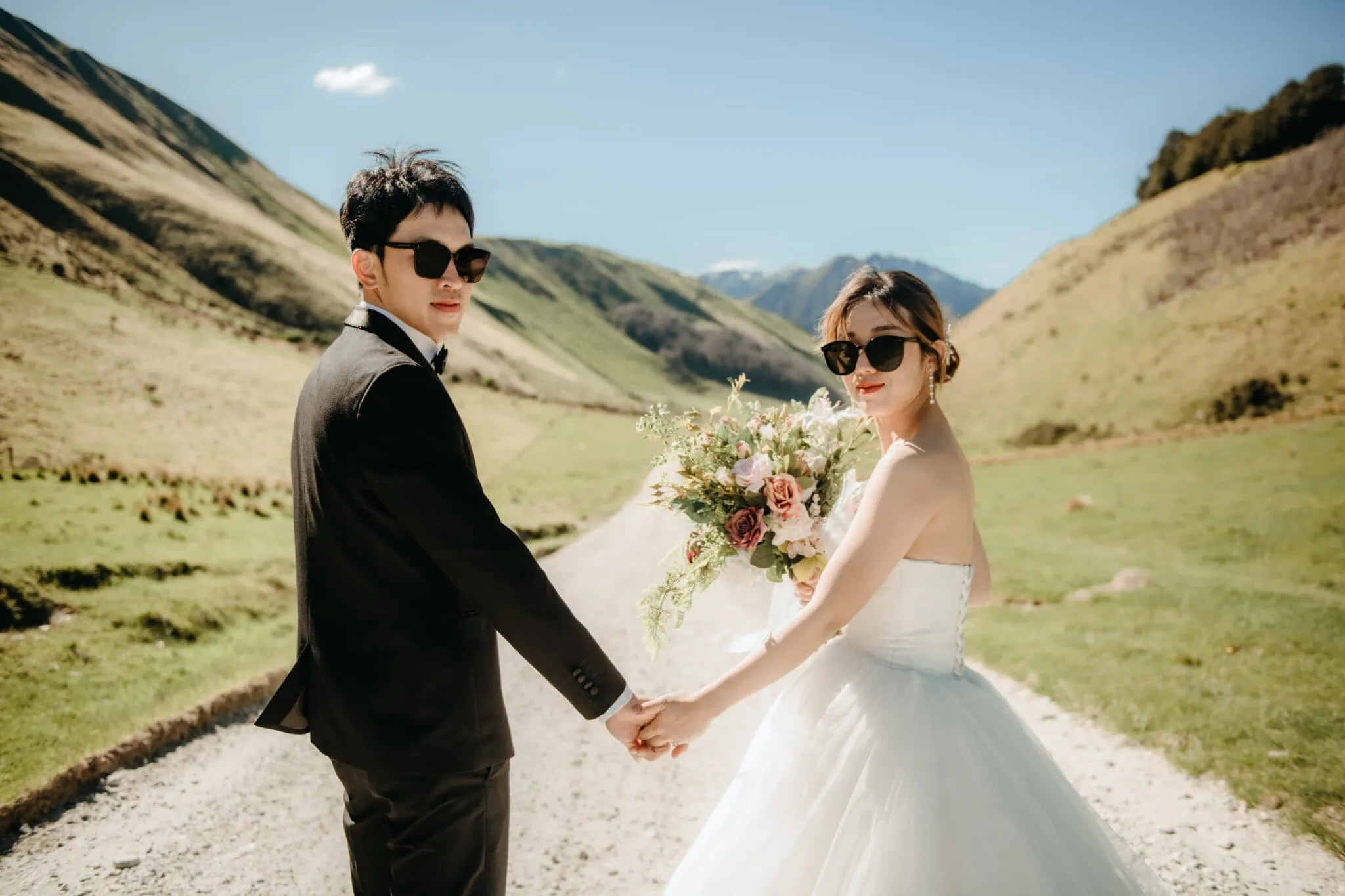 queenstown new zealand heli wedding elopement photographer videographer | A bride and groom, both wearing sunglasses, stand holding hands on a sunny, mountainous path in Queenstown. The bride holds a bouquet of flowers and they both look back at the camera, capturing the magical moment of their Heli Pre-Wedding Shoot in New Zealand.