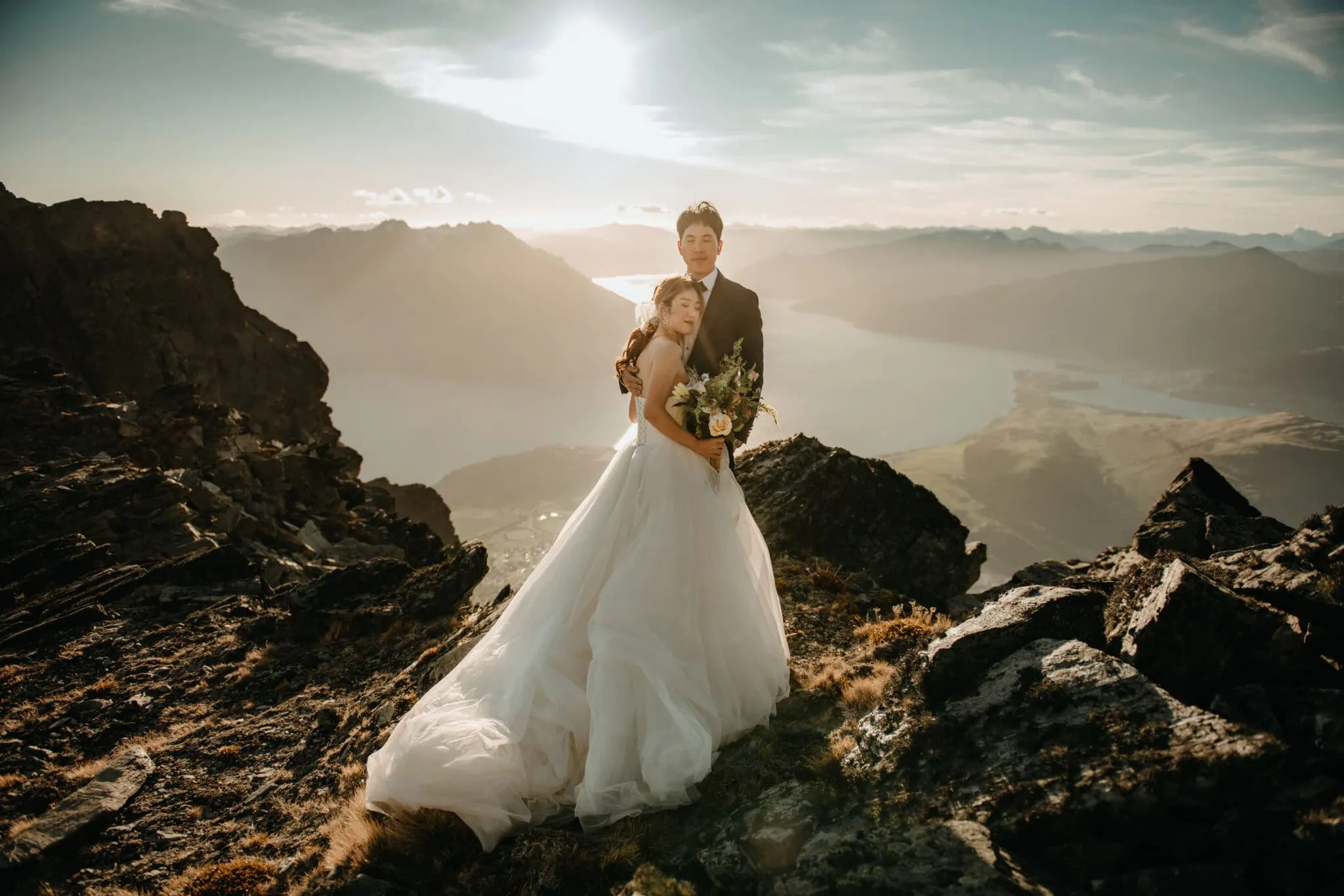 queenstown new zealand heli wedding elopement photographer videographer | Sabrina and Wilson stand together on a rocky mountain landscape in Queenstown, NZ, with a serene lake and majestic peaks in the background. The bride wears a white gown, and the groom is in a black suit, capturing their Heli Pre-Wedding moment.