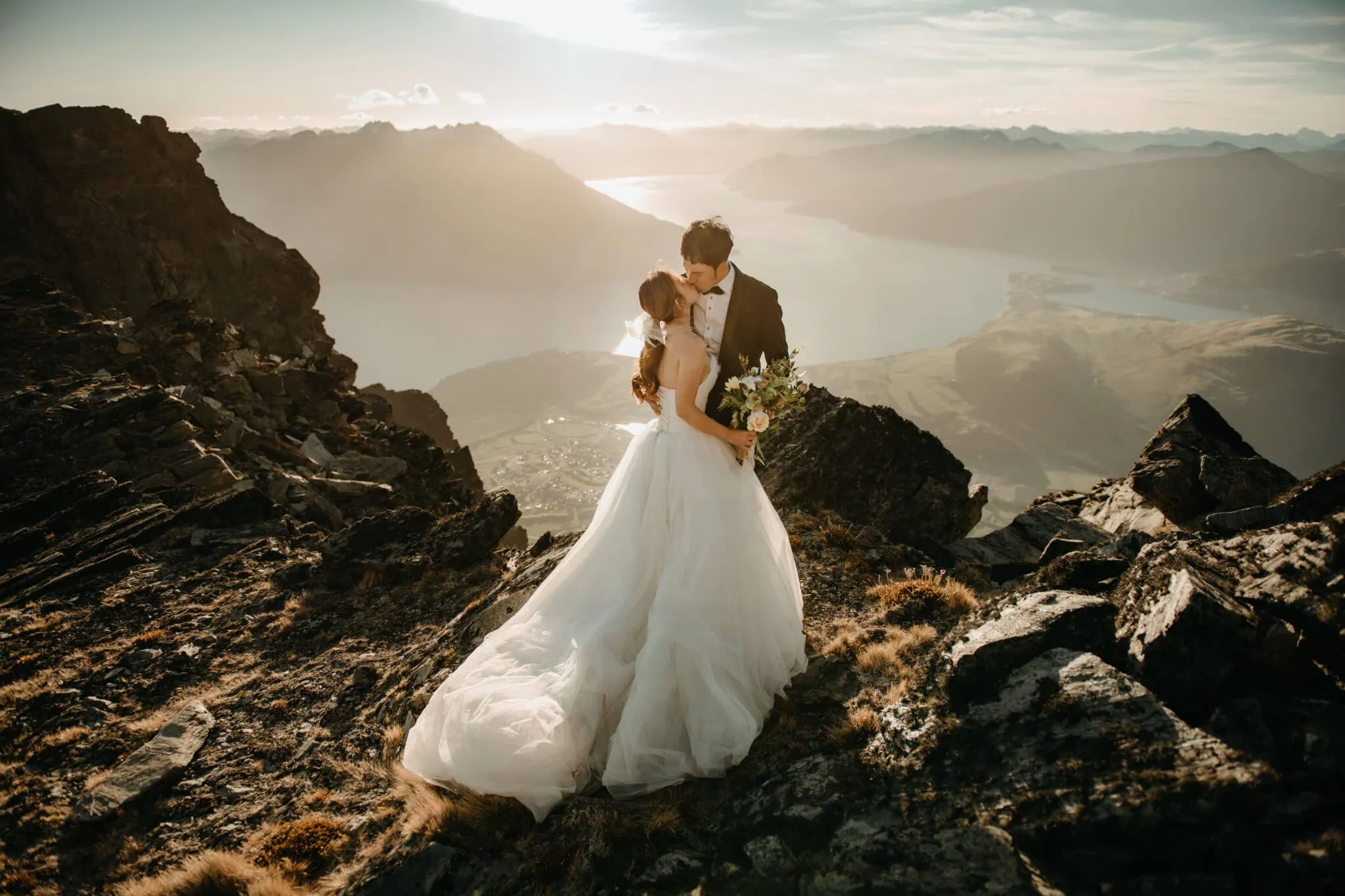 queenstown new zealand heli wedding elopement photographer videographer | A bride and groom, dressed in wedding attire, share a kiss on a rocky mountaintop overlooking a serene lake and distant mountains during sunset, capturing the magic of their NZ pre-wedding shoot in Queenstown.