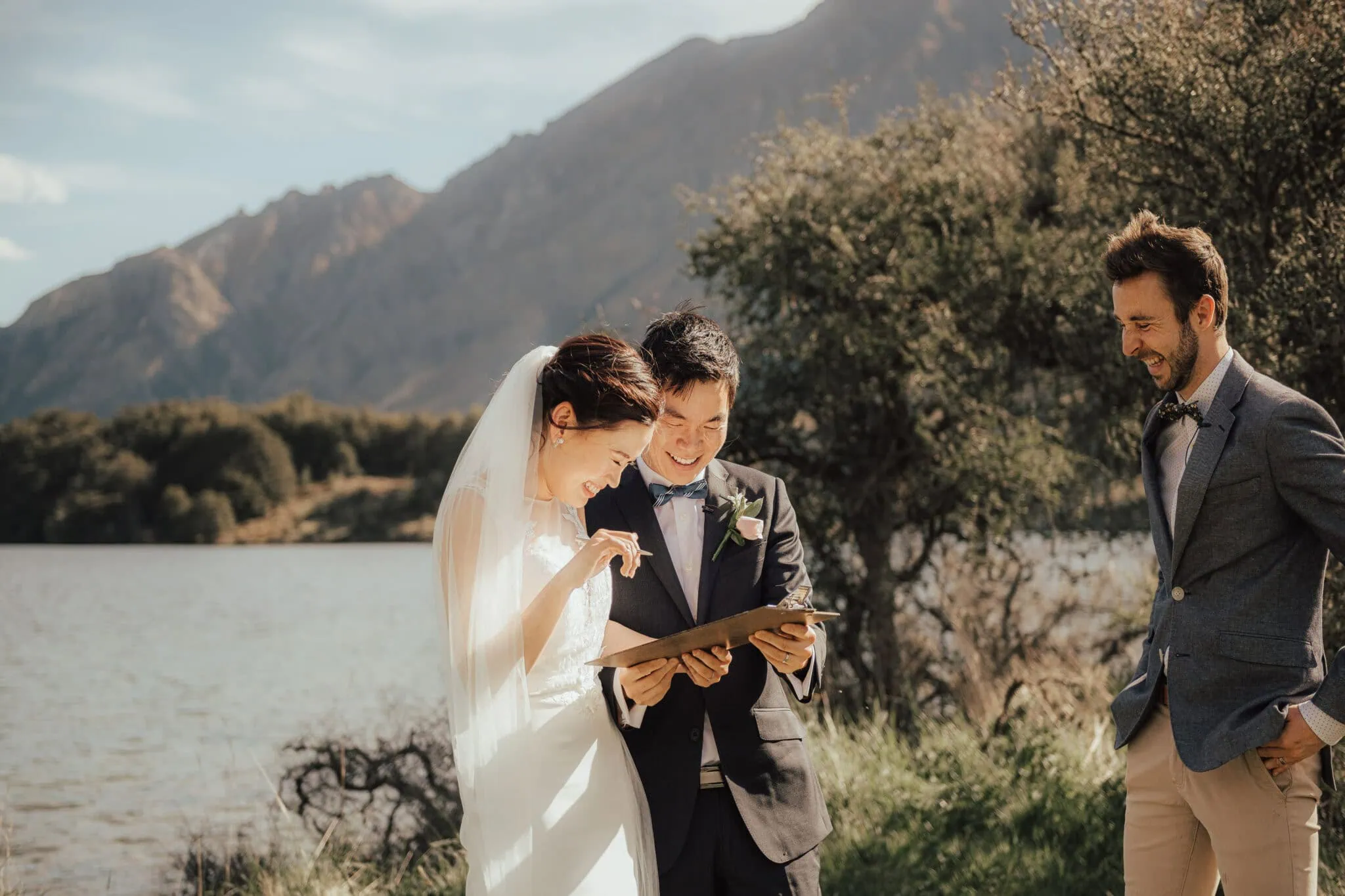 queenstown new zealand heli wedding elopement photographer videographer | Bride and groom smiling and looking at a clipboard outdoors with a lake and mountains in the background in Queenstown, NZ, accompanied by another person in a suit.