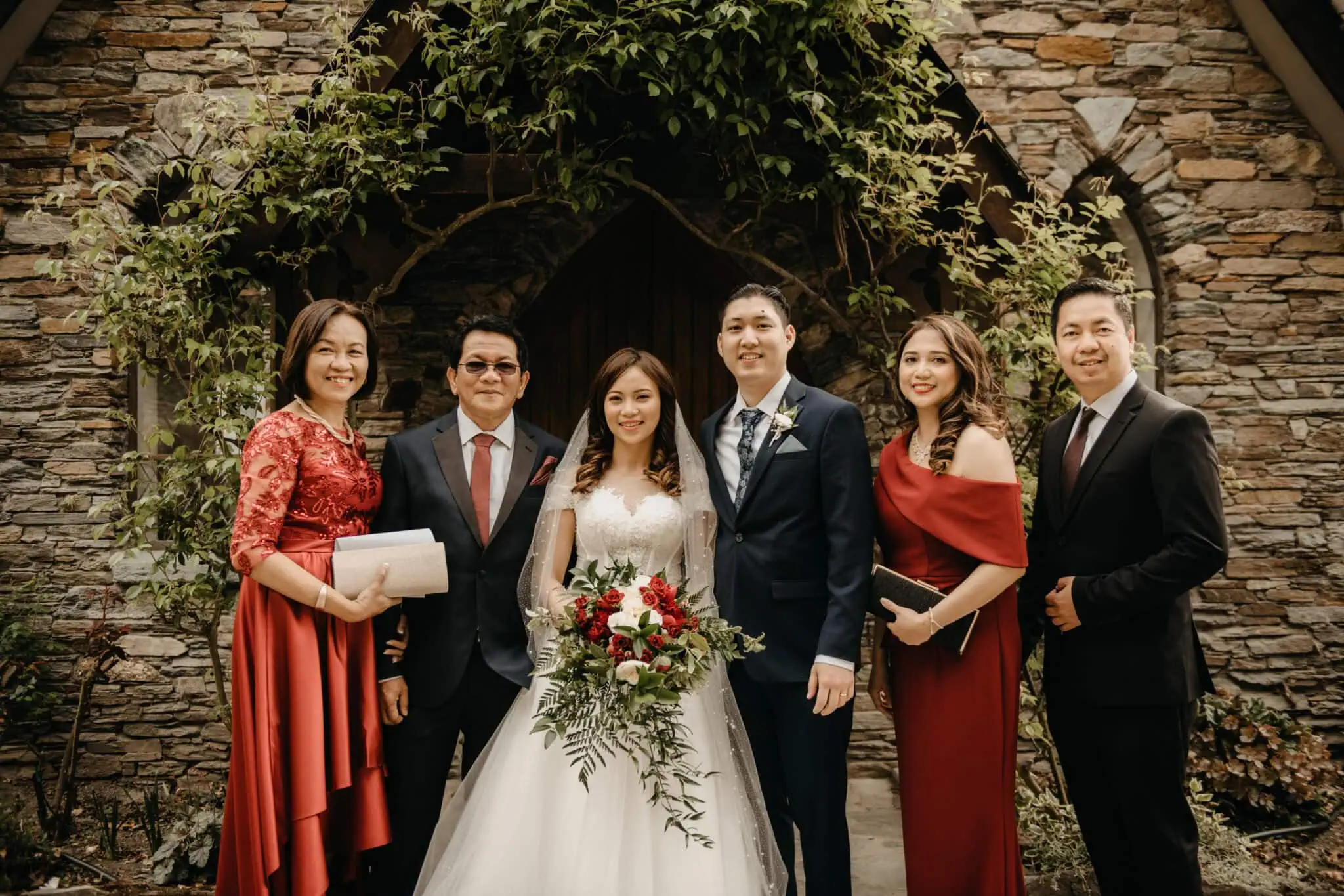 queenstown new zealand heli wedding elopement photographer videographer | A bride and groom stand with four others, dressed formally, in front of a stone building adorned with greenery at Stoneridge. Everyone is smiling and holding various items, capturing the joy of their Queenstown wedding.