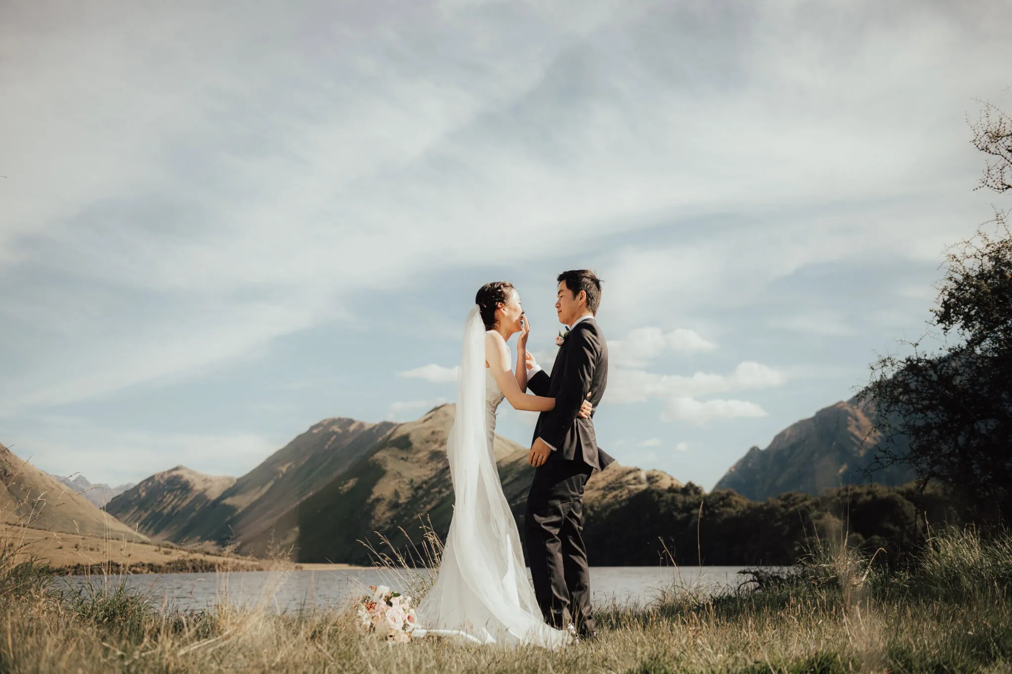 queenstown new zealand heli wedding elopement photographer videographer | Chloe & Jimmy stand close, facing each other in front of a serene lake and the majestic mountains of Queenstown under a clear sky. The bride wears a white dress and veil, while the groom is in a black suit, capturing the essence of their intimate elopement wedding.