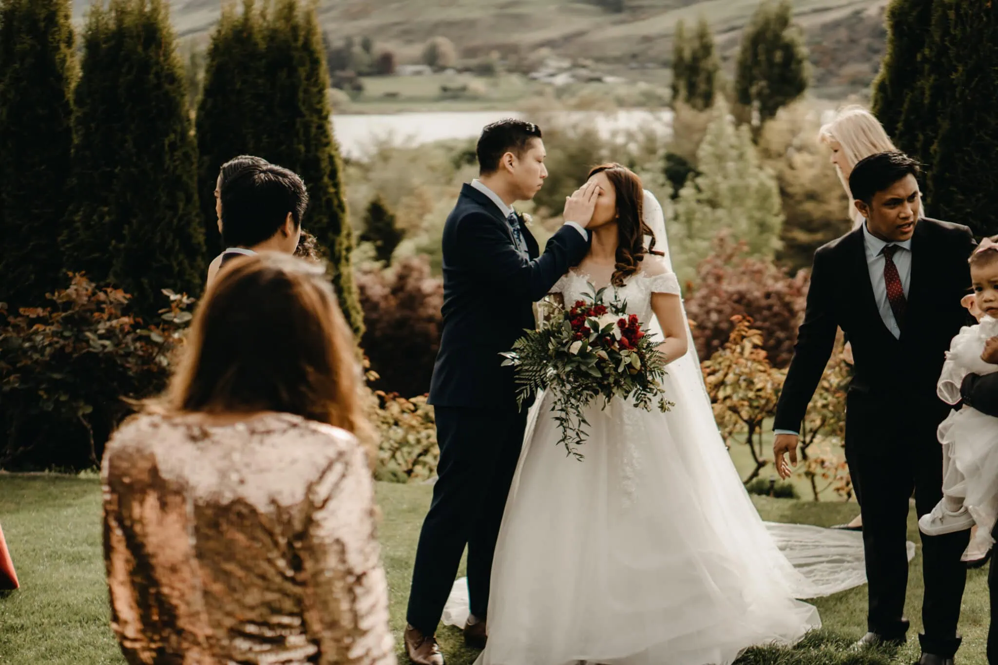 queenstown new zealand heli wedding elopement photographer videographer | A bride and groom stand in a lush outdoor setting at Stoneridge Wedding in Queenstown, with the groom touching the bride's face. The bride holds a bouquet, and several other people are around them. Trees and a lake are visible in the background, capturing the enchanting NZ landscape.