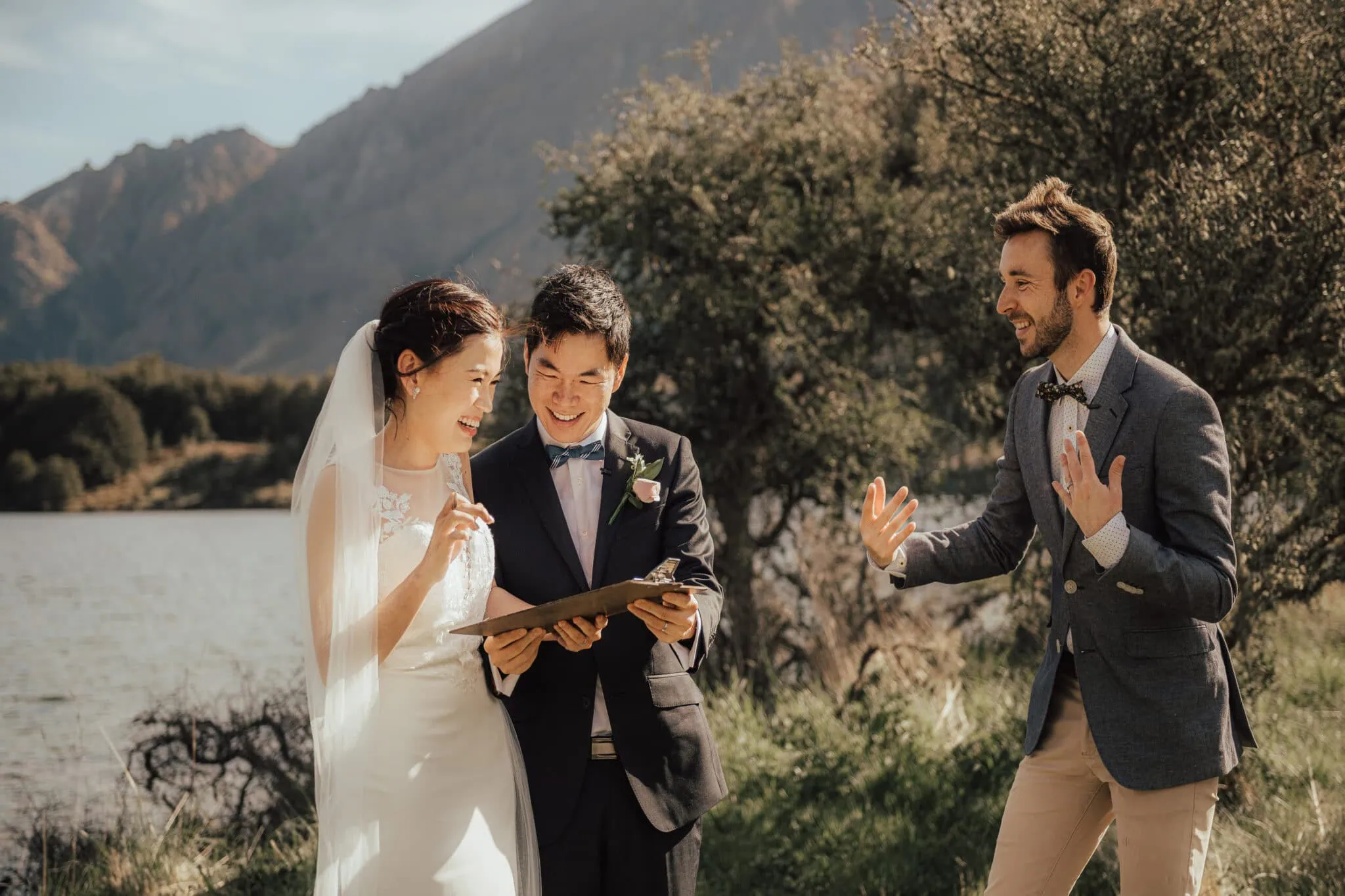 queenstown new zealand heli wedding elopement photographer videographer | A bride and groom stand outdoors by a lake in Queenstown, holding a clipboard and smiling, while a man next to them gestures animatedly. The setting captures the natural beauty of trees and mountains in the background, perfect for their intimate wedding elopement.