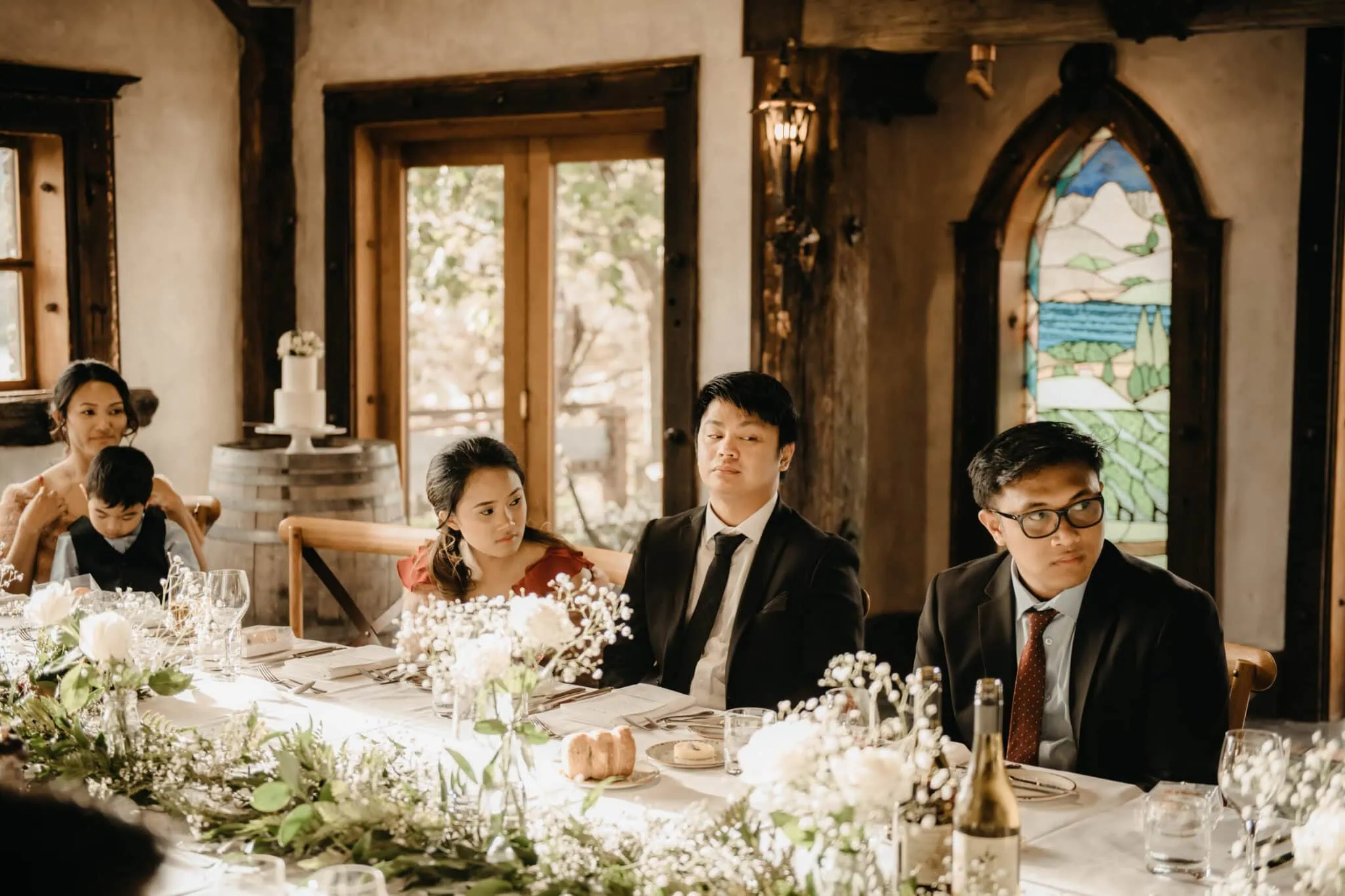 queenstown new zealand heli wedding elopement photographer videographer | A group of people in formal attire sit at a long table with a floral centerpiece, wine, and table settings in a rustic room with stained glass windows at the picturesque Stoneridge Wedding venue for Natalya & John’s celebration in Queenstown NZ.