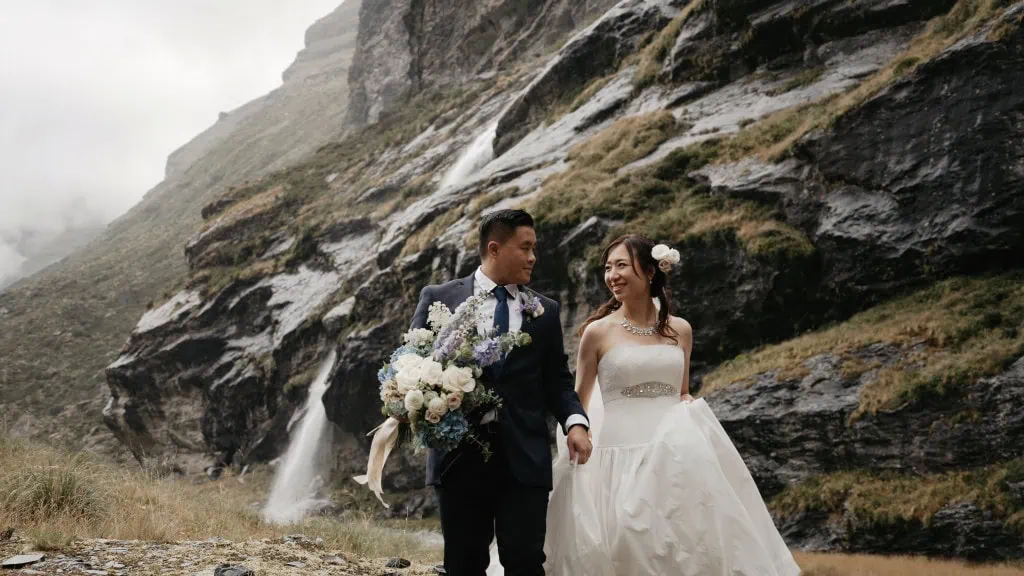 queenstown new zealand heli wedding elopement photographer videographer | Hitomi & Winston, holding hands and smiling at each other, walk outdoors in front of a rocky waterfall backdrop. The groom holds a bouquet of flowers. Their wedding takes place at the stunning Earnslaw Burn Heli-Wedding location.