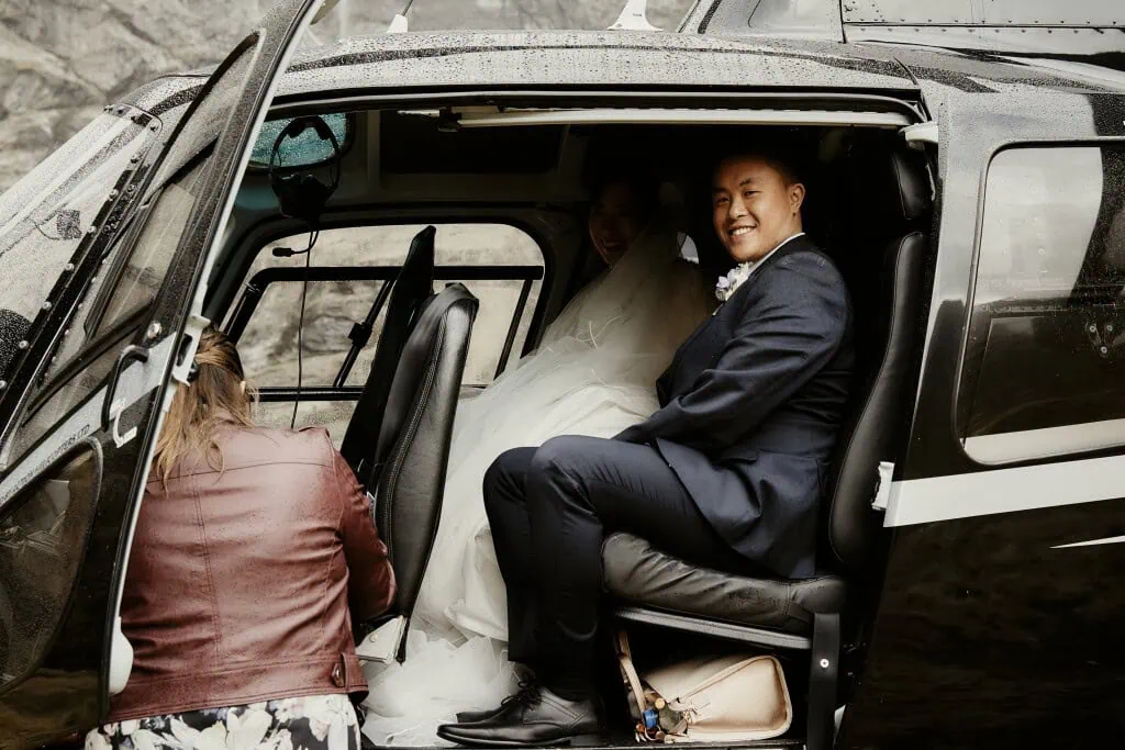 queenstown new zealand heli wedding elopement photographer videographer | Hitomi & Winston, dressed in wedding attire, sit inside a helicopter during their Earnslaw Burn Heli-Wedding. A third person kneels outside, adjusting something with the door open on this rainy day.
